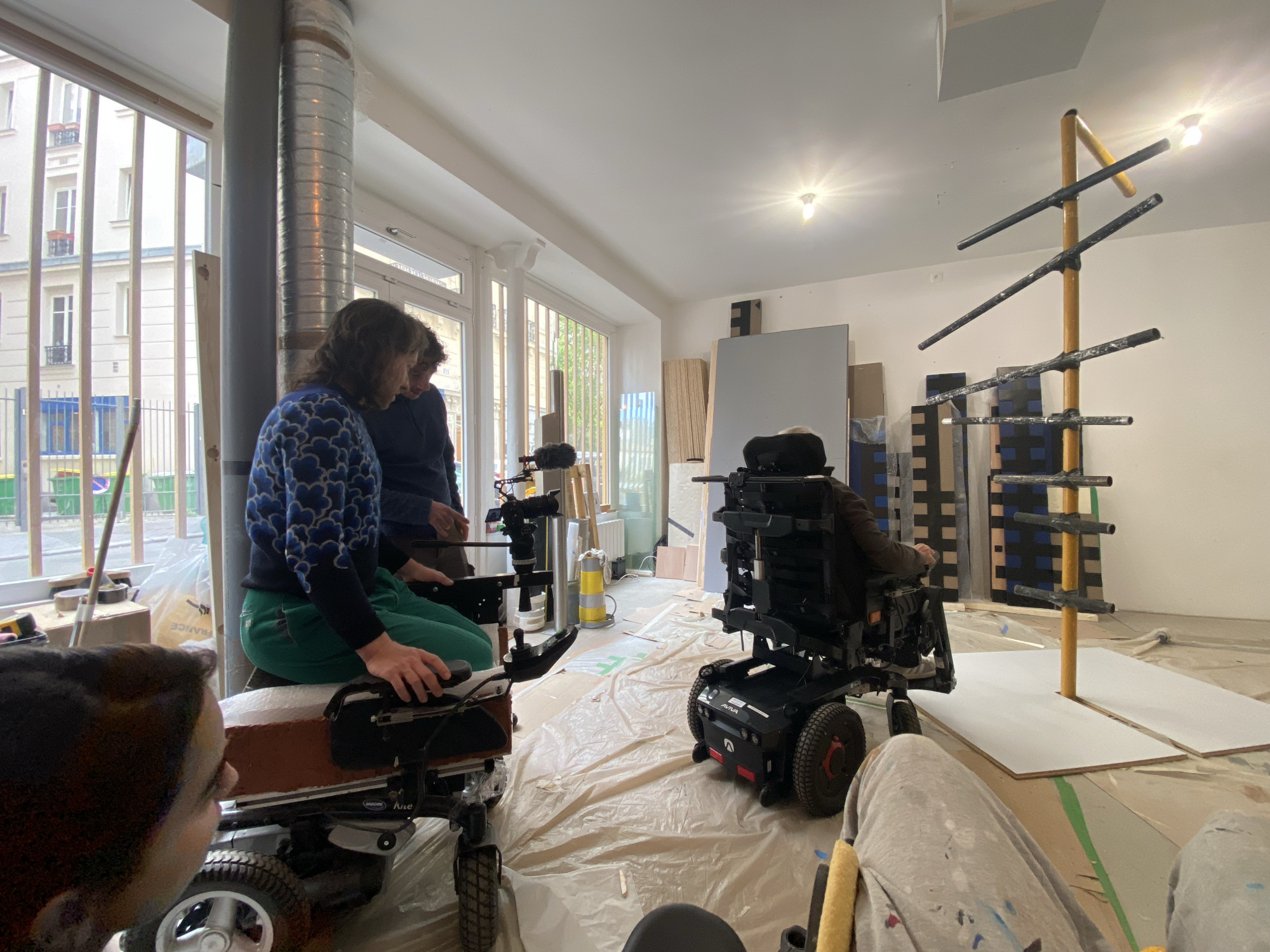 This image captures an indoor scene, likely an art studio or workshop, where a small group of people are engaged in an activity involving motorized wheelchairs and an abstract sculpture. The floor is covered with protective plastic sheeting, suggesting ongoing work or preparation.
In the foreground to the left, a person with dark, curly hair, wearing a blue patterned sweater and green pants, is seated on a brown-cushioned motorized wheelchair. They appear to be interacting with a camera and microphone setup mounted on the device. Another person stands beside them, leaning over and looking intently at the setup or something in the wheelchair operator's hands, wearing a dark top. A fourth person's head is partially visible in the far left foreground. To the right, another individual is seen from behind, seated in a black motorized wheelchair branded "AVIVA" and "Invacare," wearing light-colored pants. This person is facing the abstract sculpture.
The main object of interest in the background is a tall, golden-yellow vertical pole with several black horizontal bars attached at various heights and angles, resembling an abstract sculpture or a specialized rack. It stands on two white rectangular boards placed on the protective sheeting. Behind this sculpture, several other flat panels, some plain and some with blue and black patterns, lean against the wall, possibly other artworks or materials.
Large windows dominate the left wall, offering a view of an exterior street scene with white, multi-story buildings typical of Parisian architecture, and some greenery. A large, silver ventilation pipe runs vertically near the windows. Overhead light fixtures illuminate the space. The scene appears to be taking place during the daytime, with natural light complementing the indoor lighting. The individuals seem focused on their tasks, possibly setting up for filming, an art installation, or a technical discussion. Visible text includes "AVIVA" and "Invacare" on the black wheelchair, and "FRANCE" on a plastic bag seen through the window.