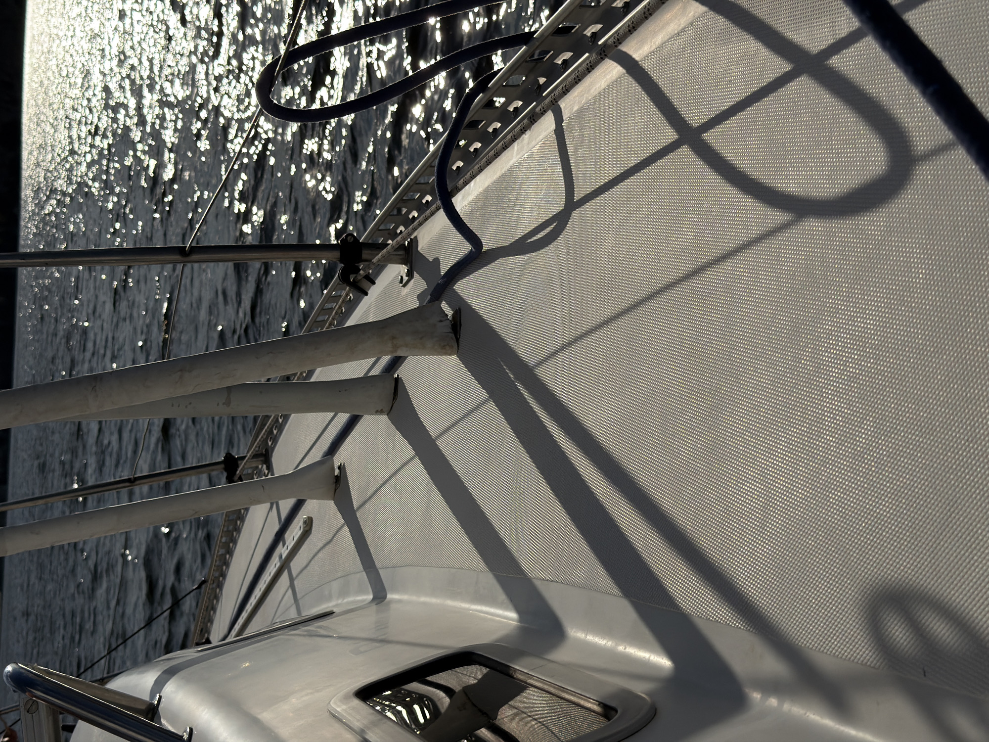 The image shows the deck of a sailboat on a body of water. The deck is covered with a textured gray material, and there are metal railings along the edge. The water is dark with shimmering reflections from the sun. The scene appears to be from a boat sailing on a river or lake, possibly the Volga River, given the reference to Kamyshin, Russia. The lighting suggests it is either morning or late afternoon. There are no people visible in the image, and no specific actions or interactions are depicted. The overall impression is one of quiet sailing or a paused moment on the water.