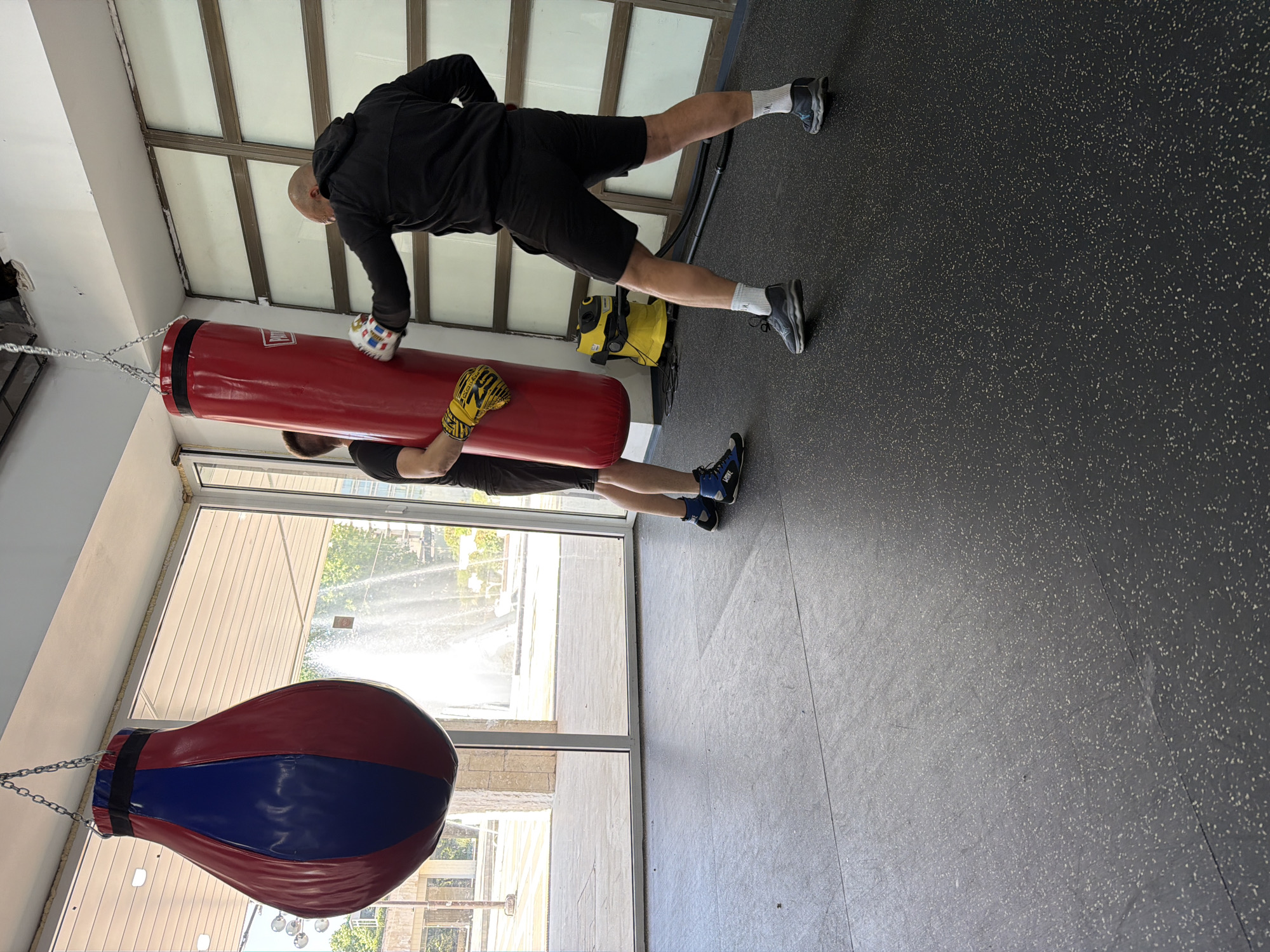 Two men are boxing in a gym in Blagoevgrad, Bulgaria. One man in black is punching a red punching bag, wearing a yellow boxing glove on his left hand and a white-patterned glove on his right. The second man is holding the bag. A second, red and blue teardrop-shaped punching bag hangs nearby. A yellow floor cleaning machine is partially visible behind the first man. Large windows are in the background. The floor is gray.