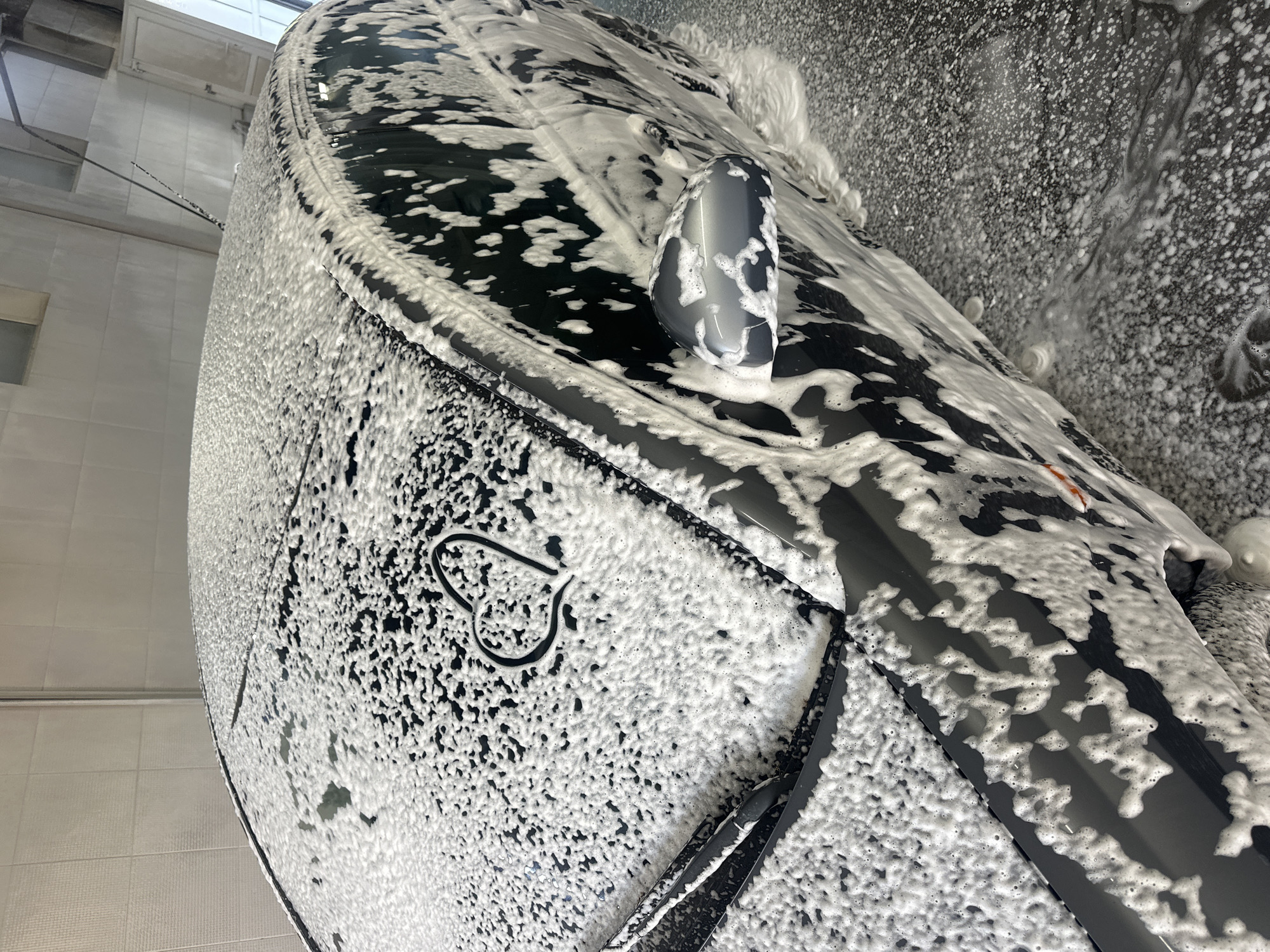 The image shows a black car covered in white soap foam. The car is likely being washed in Ternopil, Ukraine, but this is not definitively confirmed by the image. A side mirror is visible. A design resembling a heart with a downward extension is etched into the foam on the car's hood. The background includes tiled walls and potentially a ceiling fixture, suggesting an indoor setting, possibly a car wash facility.