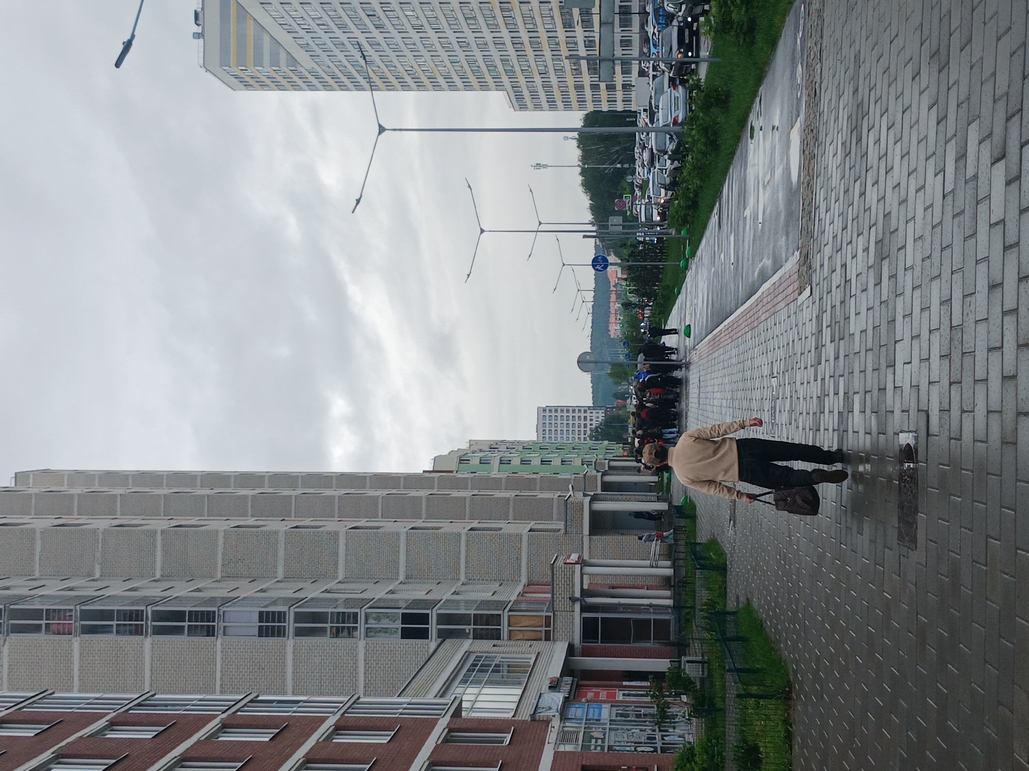 This image depicts a street scene in Yekaterinburg, Russia. On the left, a large, modern apartment building with a red-brick facade is visible. Adjacent to it is another tall apartment building, primarily light gray, with numerous windows and balconies. The street is paved with grey cobblestones, and the wet surface suggests recent rain. A sidewalk runs alongside the buildings, with a bike lane marked further out.
In the foreground, a man walks away from the camera, carrying a dark bag in his right hand. He is wearing a light-colored long-sleeved shirt and dark trousers. Beyond him, a group of people are gathered on the sidewalk, facing away from the camera, and appear to be queuing or observing something.
Further down the street, a line of cars is parked and in motion. Several lampposts are visible along the street. In the background, more apartment buildings and trees can be seen, with a cloudy sky overhead. The scene appears to be in a residential area of the city.
