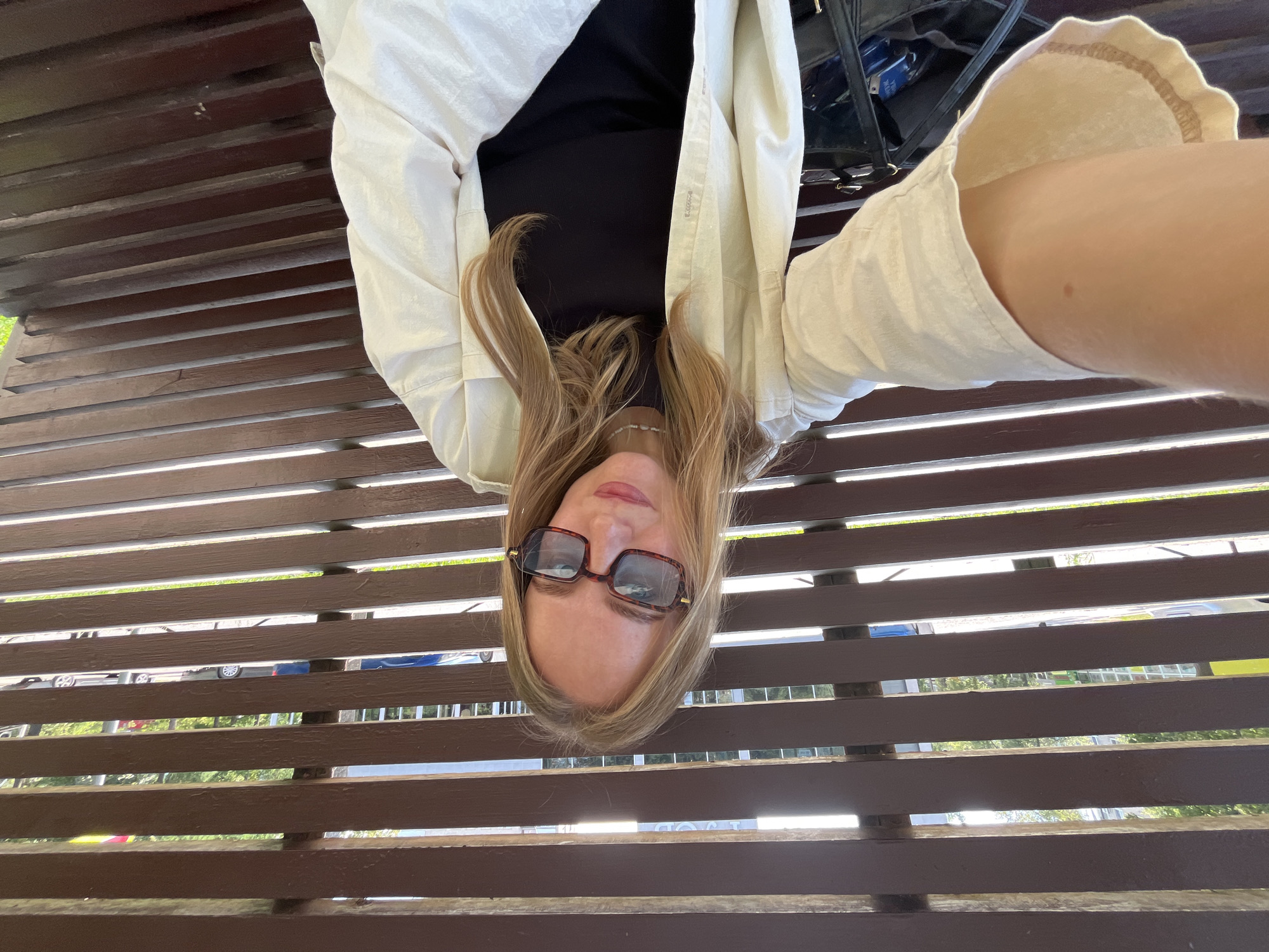The image shows a young woman sitting on a wooden slatted bench outdoors. She is wearing a black top and a light-colored open jacket. She has blonde hair and is wearing large, tortoiseshell-patterned glasses. Her face is tilted towards the camera, and she is looking directly at it. Her expression is neutral.
The setting appears to be a park or public space, with trees and greenery visible in the background through the slats of the bench. Sunlight is filtering through, suggesting it is daytime. The bench is made of dark brown wood and is oriented horizontally in the frame. Behind the woman, a black bag is visible, partially obscured by her jacket. The image is taken from a low angle and is rotated, so the woman appears upside down.
No visible text is present in the image. The woman's attire and the lighting suggest a casual daytime activity, possibly a break or a moment of relaxation. The overall mood is relaxed and natural. Given the provided location context of Chaikovsky, Russia, the scene is likely set in a Russian public space.