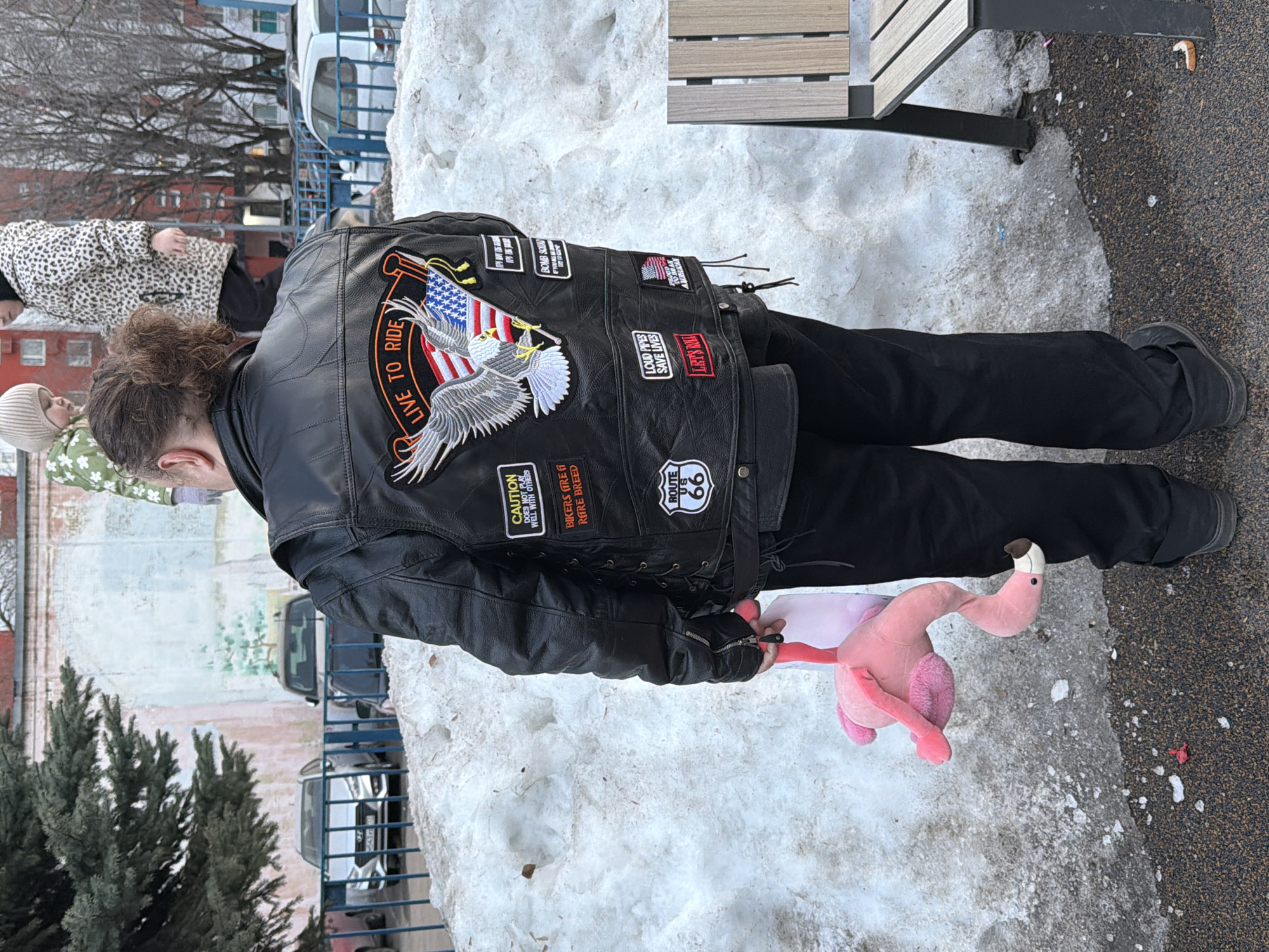 An adult, viewed from behind, stands outdoors in a snow-covered urban environment in Khimki, Russia. The individual has brown, curly hair tied back in a bun and is wearing a black leather vest over a dark garment, paired with black pants and dark shoes. The vest is extensively decorated with patches, including a prominent central patch featuring an American bald eagle over an American flag with a banner reading "LIVE TO RIDE." Other visible patches include "CAUTION DOES NOT PLAY WELL WITH OTHERS," "BIKERS ARE A RARE BREED," "ROUTE US 66," "LOUD PIPES SAVE LIVES," "LET'S ROLL!," a small American flag, and partial views of "IF IT'S HOT THE TOP IT'S THE BOOBS," "BOMB SQUAD IT'S ABOUT TIME I BLEW UP AND GOT TO RIDE," and "I'M A PROUD AMERICAN." The person holds a pink plush flamingo toy by one leg in their left hand.
The ground consists of a dark, granular surface interspersed with dirty, melting snow patches. A segment of a wooden bench with dark metal supports is visible on the right. In the mid-ground, a large mound of melting snow separates the foreground from the background elements. Beyond the snow, a light blue metal fence encloses a parking area with several parked cars, including a silver Hyundai sedan. Behind the vehicles, a brick wall features a faded mural depicting a light blue sky and a green tree silhouette. A dark green coniferous tree is partially visible on the left. In the far background, two children are positioned atop the snow mound; one child wears a light-colored beanie and a green patterned jacket, while the other wears a leopard-print jacket. Multi-story residential buildings and bare deciduous trees are visible in the distant background.