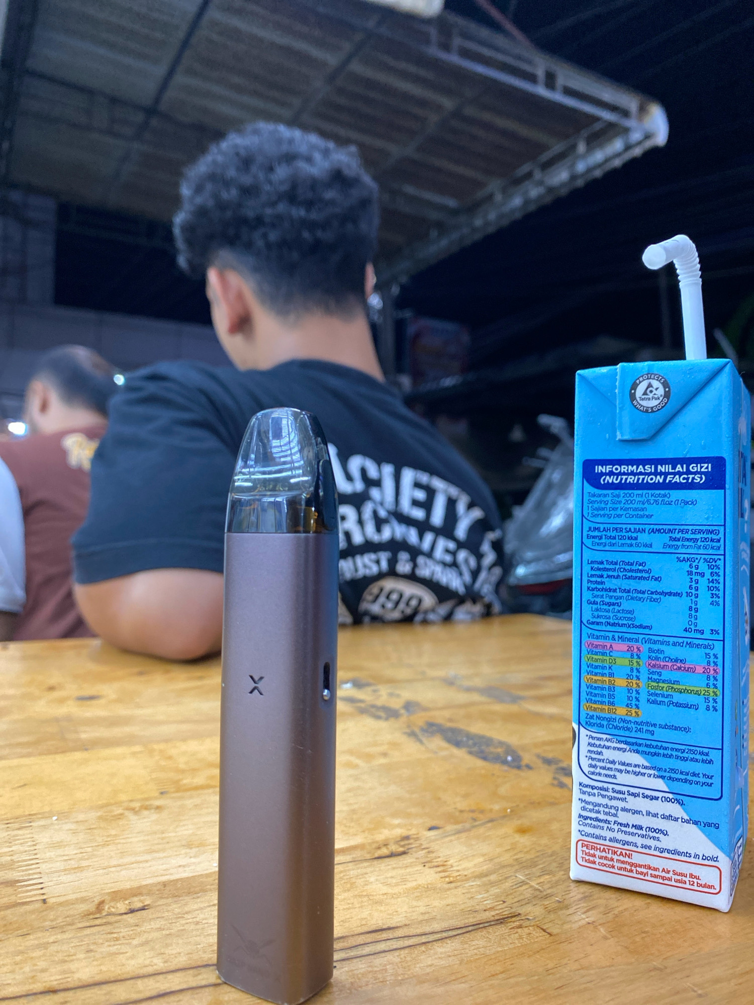 This image captures a casual moment outdoors, likely at a food stall or outdoor dining area in Pantegajah, Indonesia, indicated by the warm wooden table and the rustic ceiling structure overhead. In the foreground, a sleek, bronze-colored vape pen with a clear plastic top stands vertically on the table. To the right, a blue Tetra Pak carton of milk, featuring a white straw, is positioned. The carton displays nutritional information and warnings in Indonesian and English. In the background, two individuals are visible. One, a person with curly dark hair wearing a black t-shirt, is facing away from the camera, engrossed in conversation or activity. Another person, partially obscured, is seated to the left. The overall ambiance suggests a relaxed, informal setting.