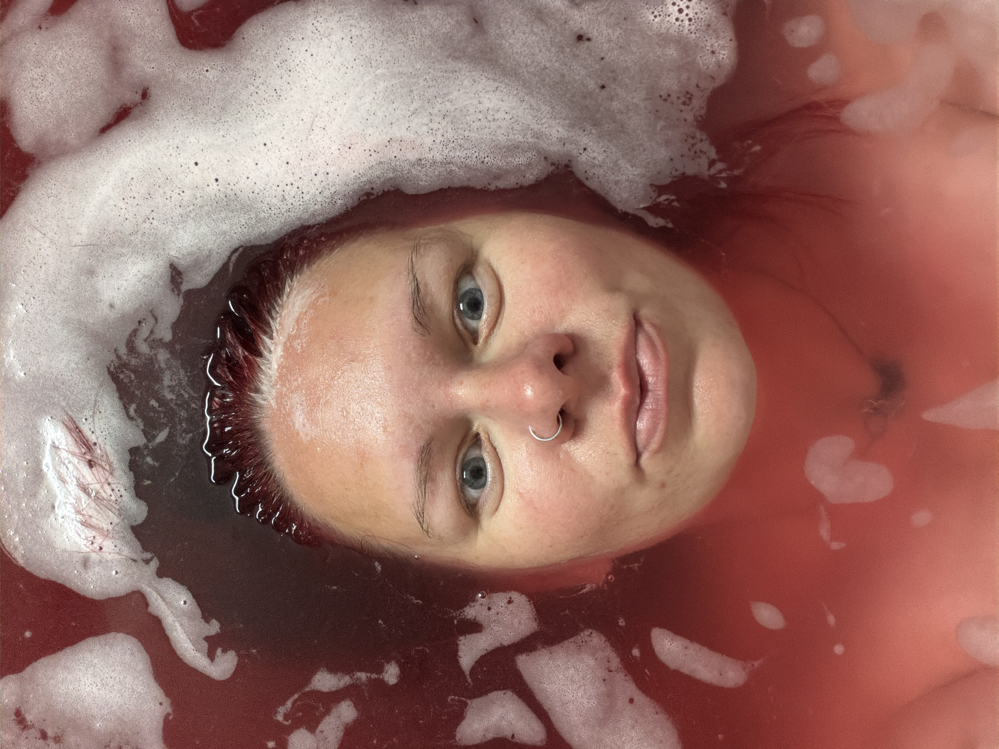 A woman lies in a bath filled with red-tinted water and white bubbles. Her face is visible above the water line, featuring blue eyes and a nose ring. Her hair, appearing dark, is partially submerged. The environment suggests a bathroom setting. Location data for Chaikovsky, Russia is not evident in the image.