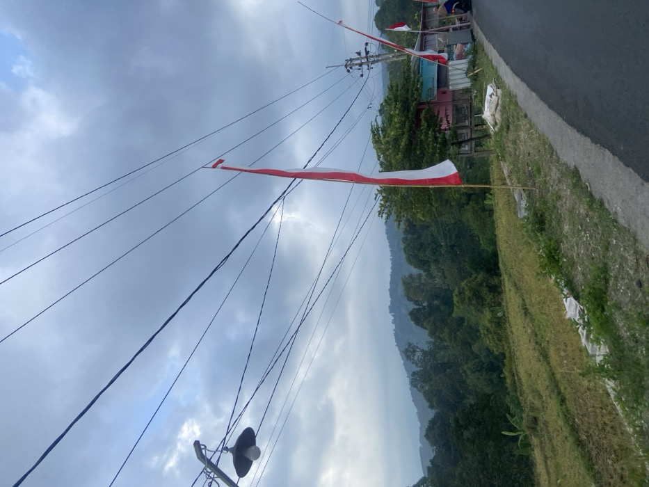 The image depicts an outdoor scene in Sukoreno, Indonesia, under an overcast sky. A dark asphalt road curves from the lower right foreground towards the upper right. To the right of the road, a dirt and grass shoulder contains multiple white sacks and two red and white Indonesian flags mounted on thin poles. The primary flag is positioned in the mid-ground, with a smaller flag visible further back. Behind these flags, a single-story structure with faded pink walls and a light blue sign or awning is present, adjacent to a wooden utility pole supporting numerous overhead electrical wires. A dark object, consistent with a motorcycle, is partially visible near the building. To the left of the road, a terraced field, appearing as cultivated land with stubble, slopes downwards towards a dense forest. In the distant background, vegetated mountains are visible beneath the cloudy sky. In the upper left, a dark streetlamp with a visible white bulb hangs from a metal arm, with multiple black electrical wires extending horizontally across the frame. No human figures are discernible.