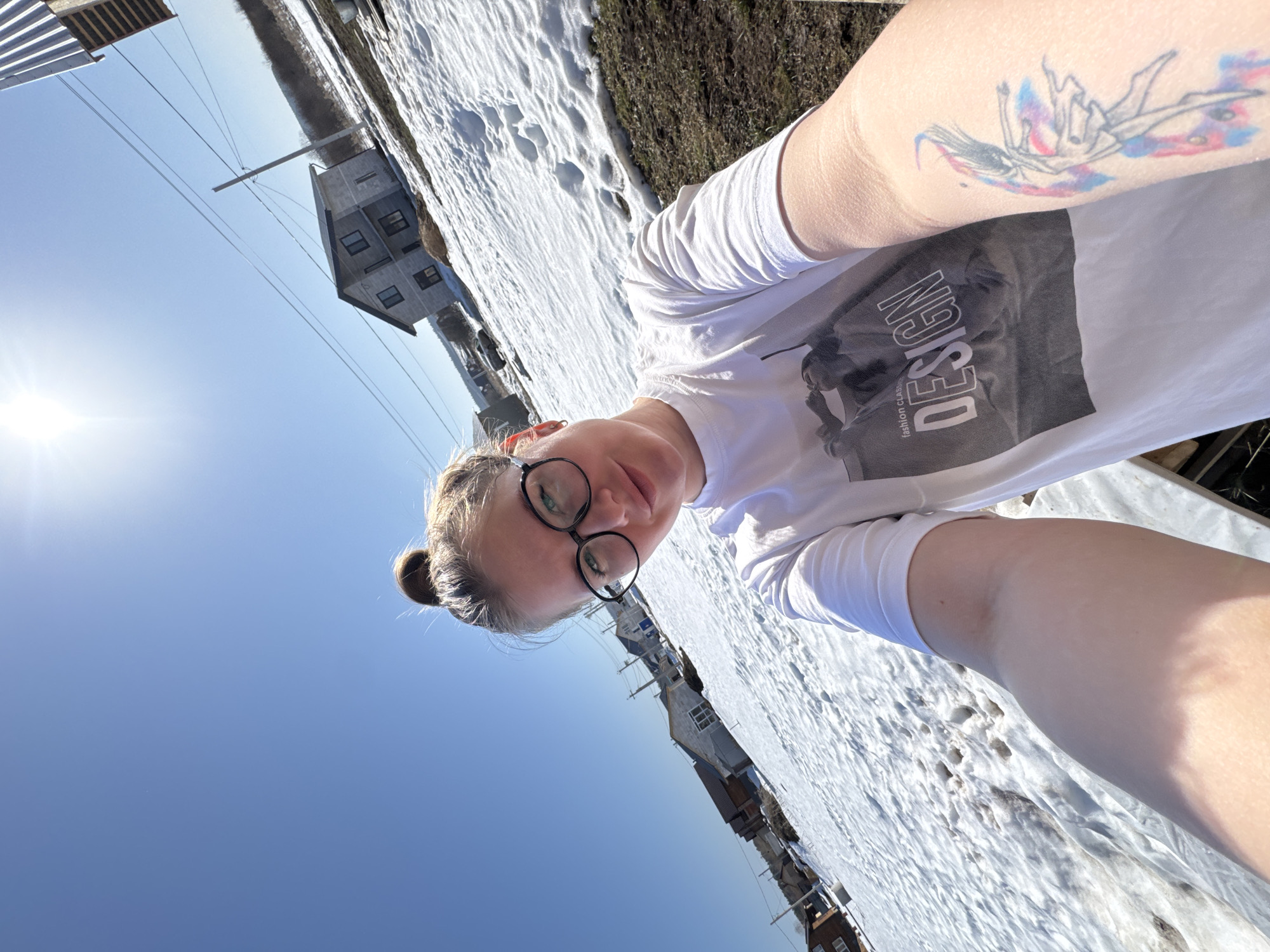 A woman is taking a selfie outdoors. She is wearing a white t-shirt with the word "DESIGN" printed on it, and round black glasses. She has a tattoo on her upper left arm. The background shows a snow-covered landscape with a few buildings, a bright blue sky, and the sun. Based on the presence of buildings and snow cover, the location is likely Gornyi (Dvurechenskoe S/P), Russia.