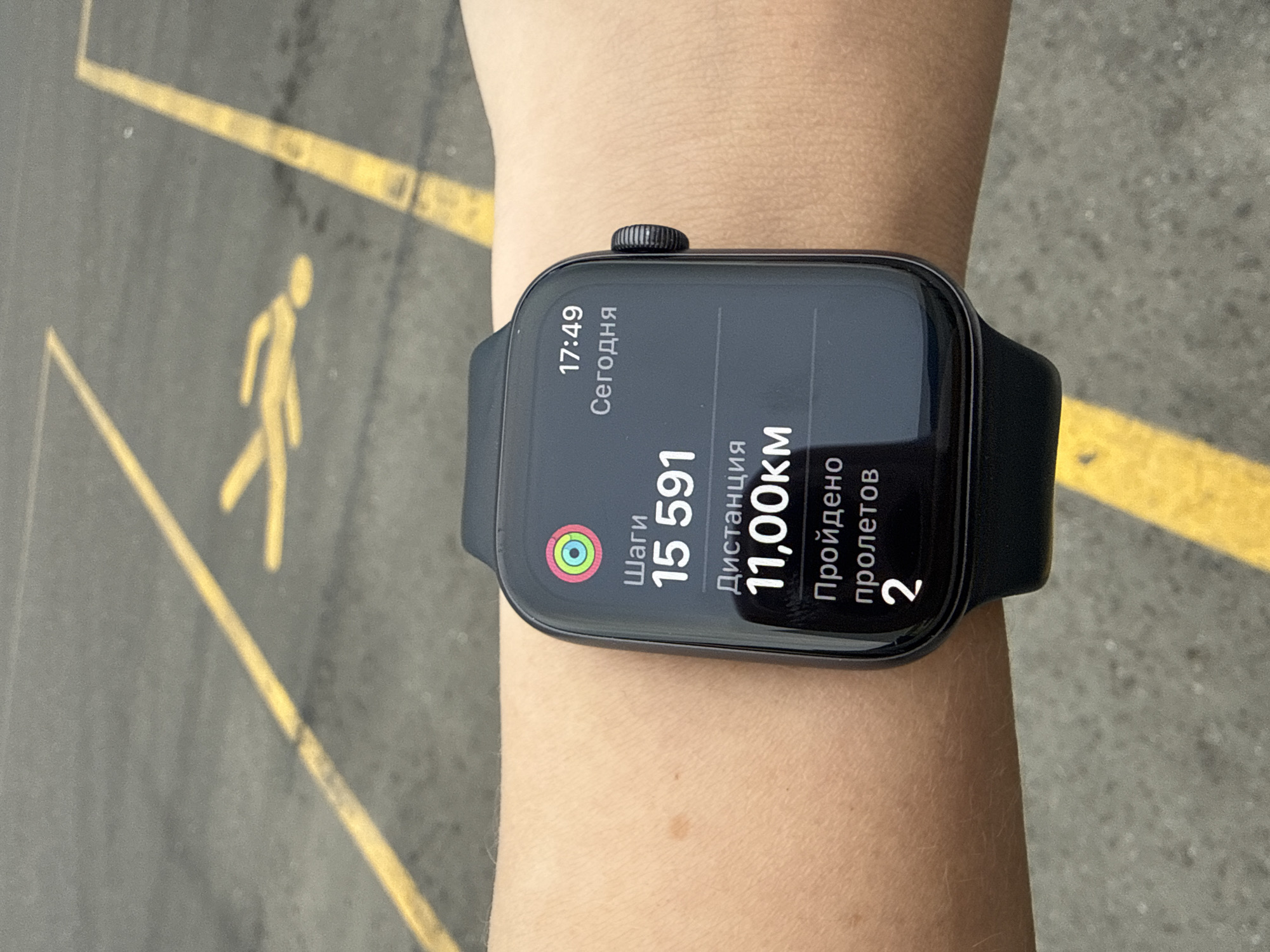 A person is wearing a black Apple Watch on their left wrist, displaying the time as 17:49 and the following statistics: 15,591 steps taken, 11.00 kilometers distance traveled, and 2 flights climbed. The background shows a gray surface with yellow pedestrian crossing markings. The scene is potentially in Lupanovo, Russia, given the user's prompt.