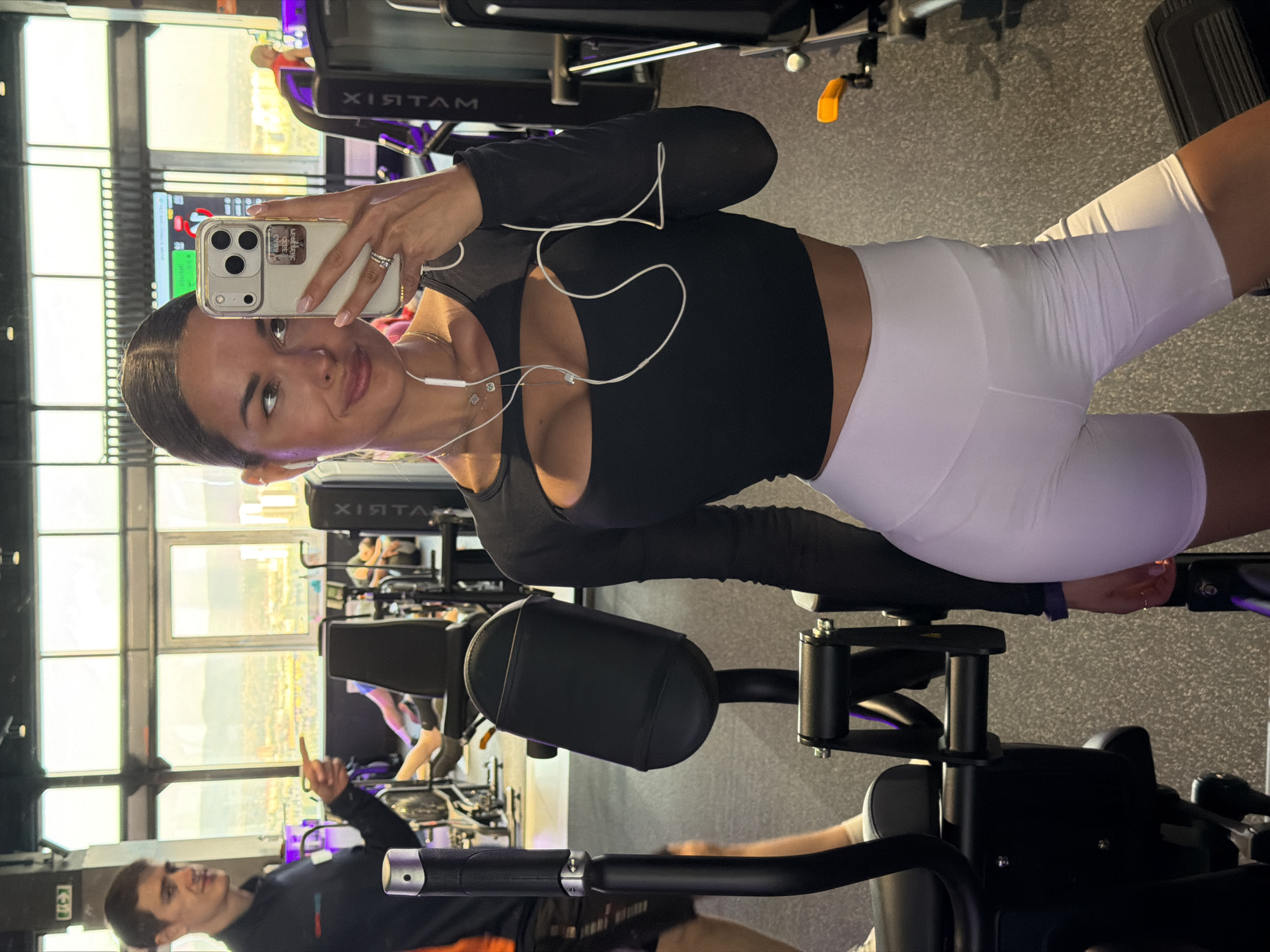 A young woman in a black long-sleeved top and white shorts is taking a selfie in a gym. She is holding a white iPhone with a case. The gym appears modern with large windows and gym equipment. A man wearing a dark shirt and jeans is in the background, pointing upward. It is impossible to verify whether the location is Krasnoyarsk, Russia.