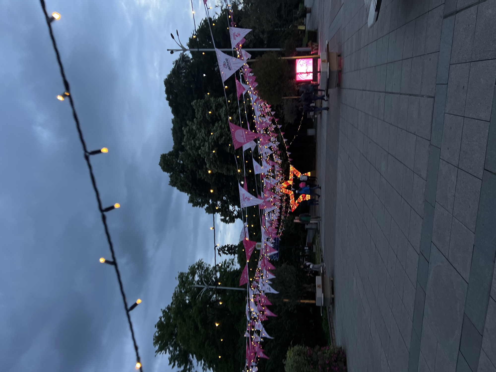 The image depicts a festive street scene in Plovdiv, Bulgaria. A pedestrian walkway paved with rectangular gray stones stretches into the distance. The sky is overcast with a dark, cloudy sky. Strings of small, warm-toned lights are strung across the walkway, along with rows of pink and white pennant flags. A large, illuminated star is situated near the end of the walkway. People can be seen walking and gathered near the illuminated star. Trees line the sides of the walkway, and a digital display emitting a pink light is visible further down the street. The overall impression is of a public space decorated for an event or holiday.