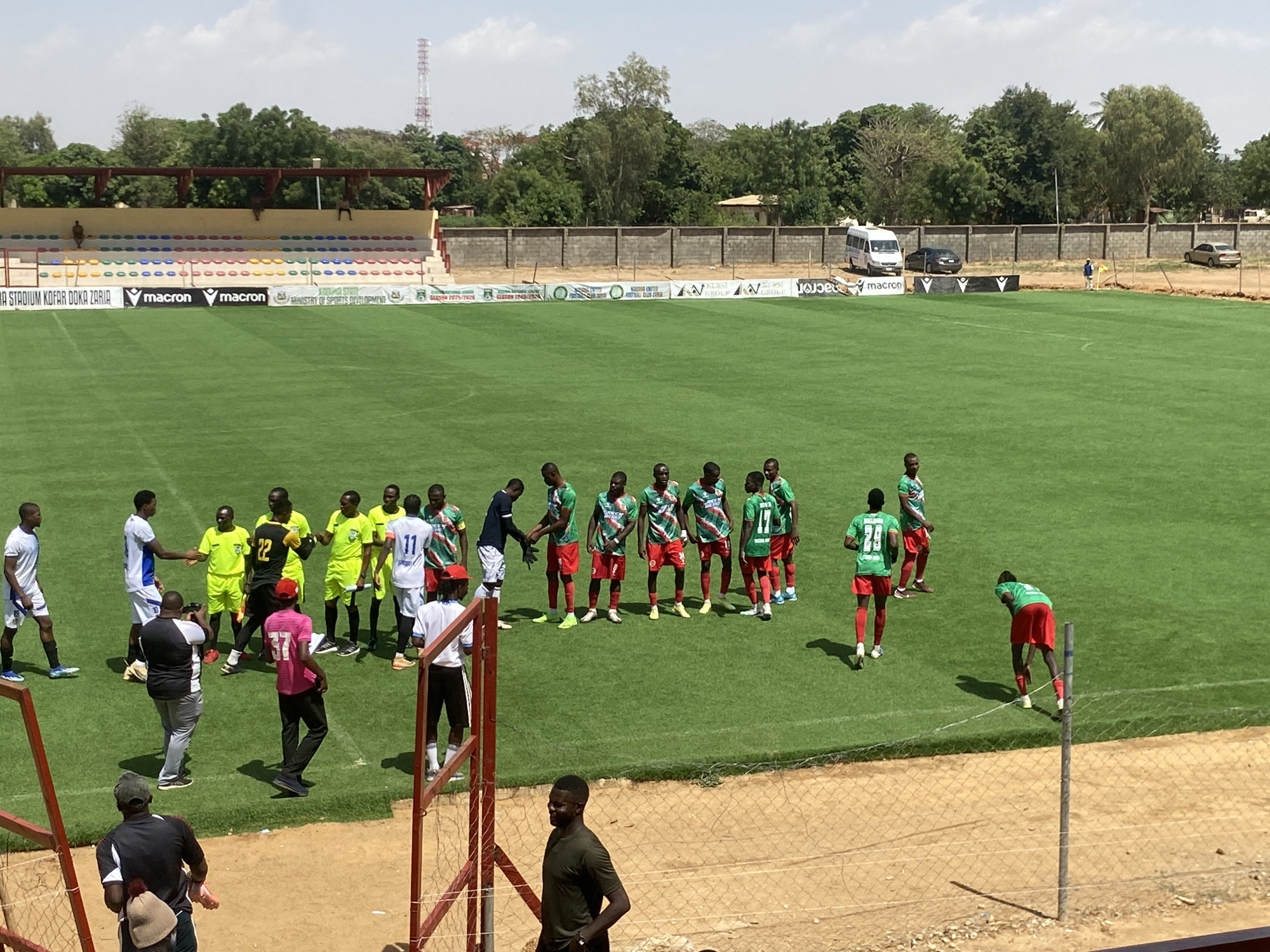 The image shows a soccer field with two teams lining up for what appears to be a match. One team is wearing white and blue uniforms, while the other is in green and red. A group of people, including a photographer and officials, are standing near the teams. Behind the field, there is a stadium with colorful seats. A wall and some parked vehicles are visible in the background. The location is Kofar Doka Stadium in Zaria, Nigeria, as indicated by the signage in the stadium.