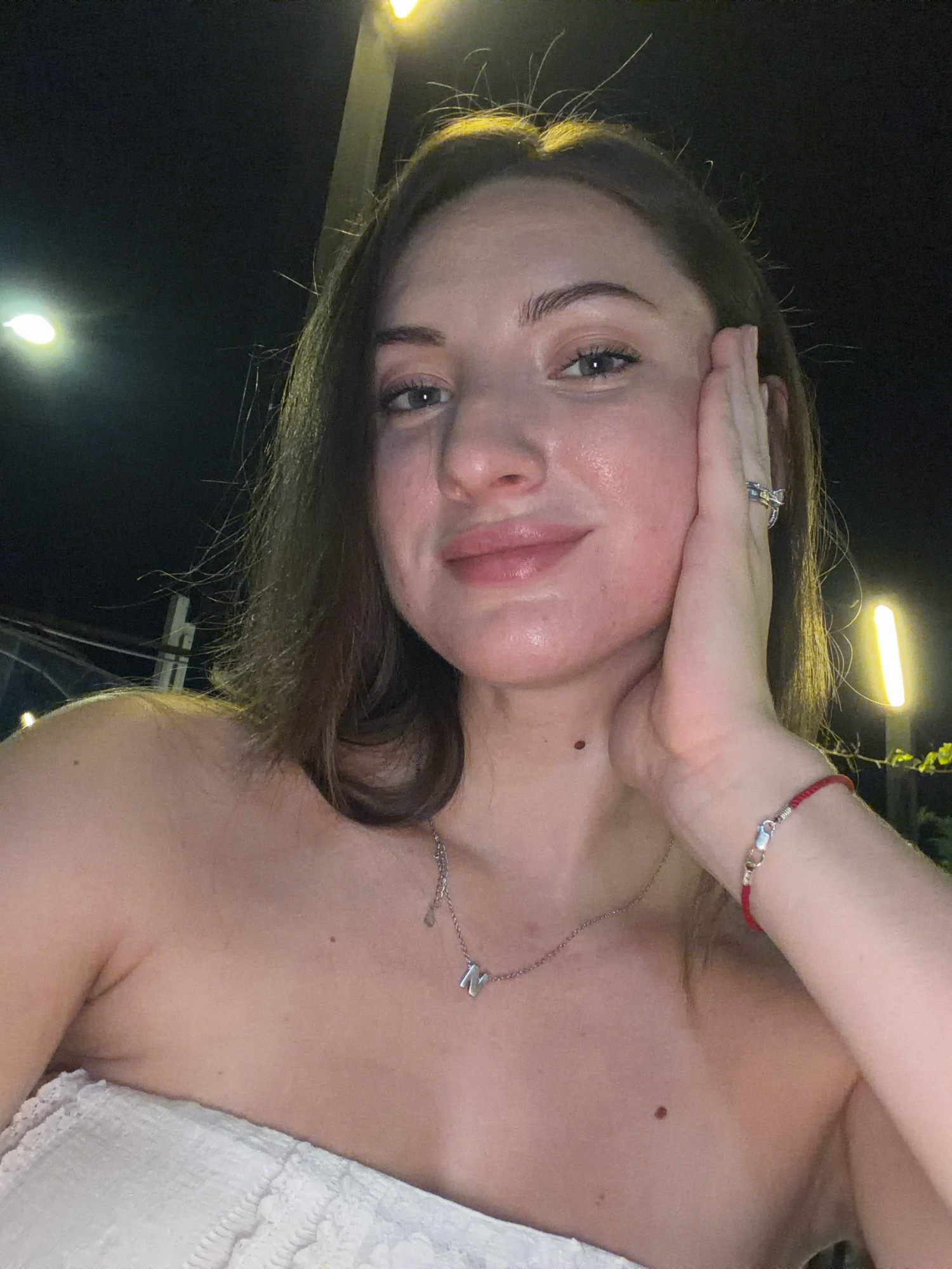 A young woman with light skin and brown hair is captured in a close-up, selfie-style photograph taken at night. She is wearing a strapless white top and a silver necklace with a letter charm. Her right hand is resting on her cheek, and she is wearing a ring on her right hand and a red string bracelet on her right wrist. Her left shoulder and upper chest are visible, revealing a mole on her chest. The background is dark, with several out-of-focus lights, suggesting an outdoor urban setting. One of these lights appears to be a street lamp. The image was taken in Sharm El Sheikh, Egypt.