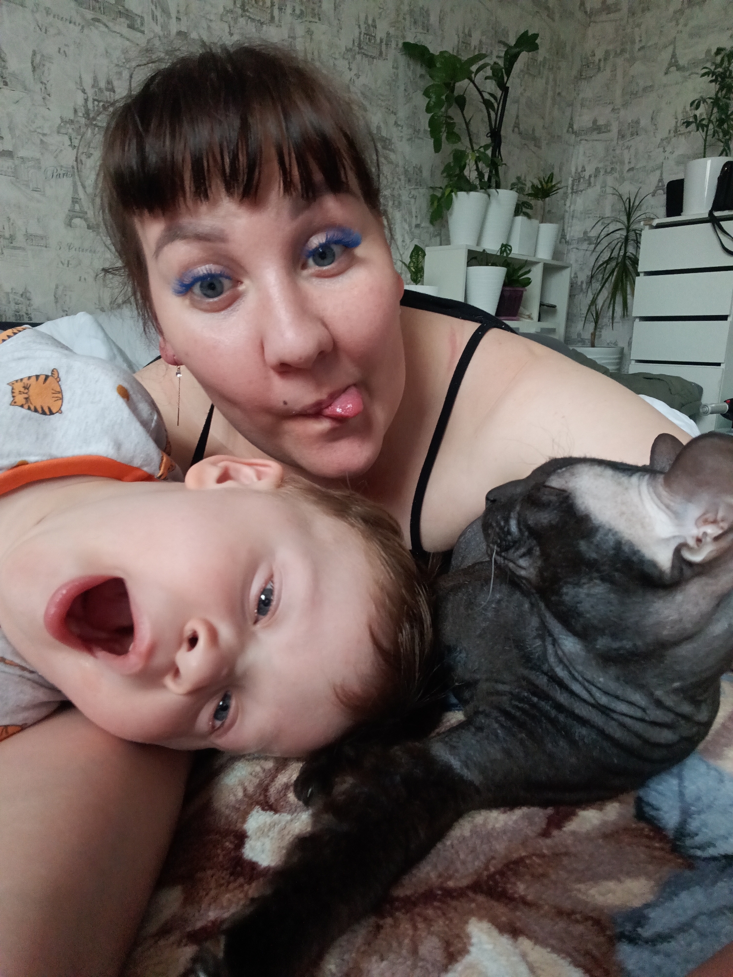 A woman, a child, and a Sphynx cat are lying on a bed. The woman has brown hair, blue eyelashes and is sticking her tongue out. The child is on their side with their mouth open. The cat is close to the child. The background includes plants in white pots on a white shelf, a white dresser, and wallpaper with an Eiffel Tower pattern. No evidence suggests a location in Osa, Russia.