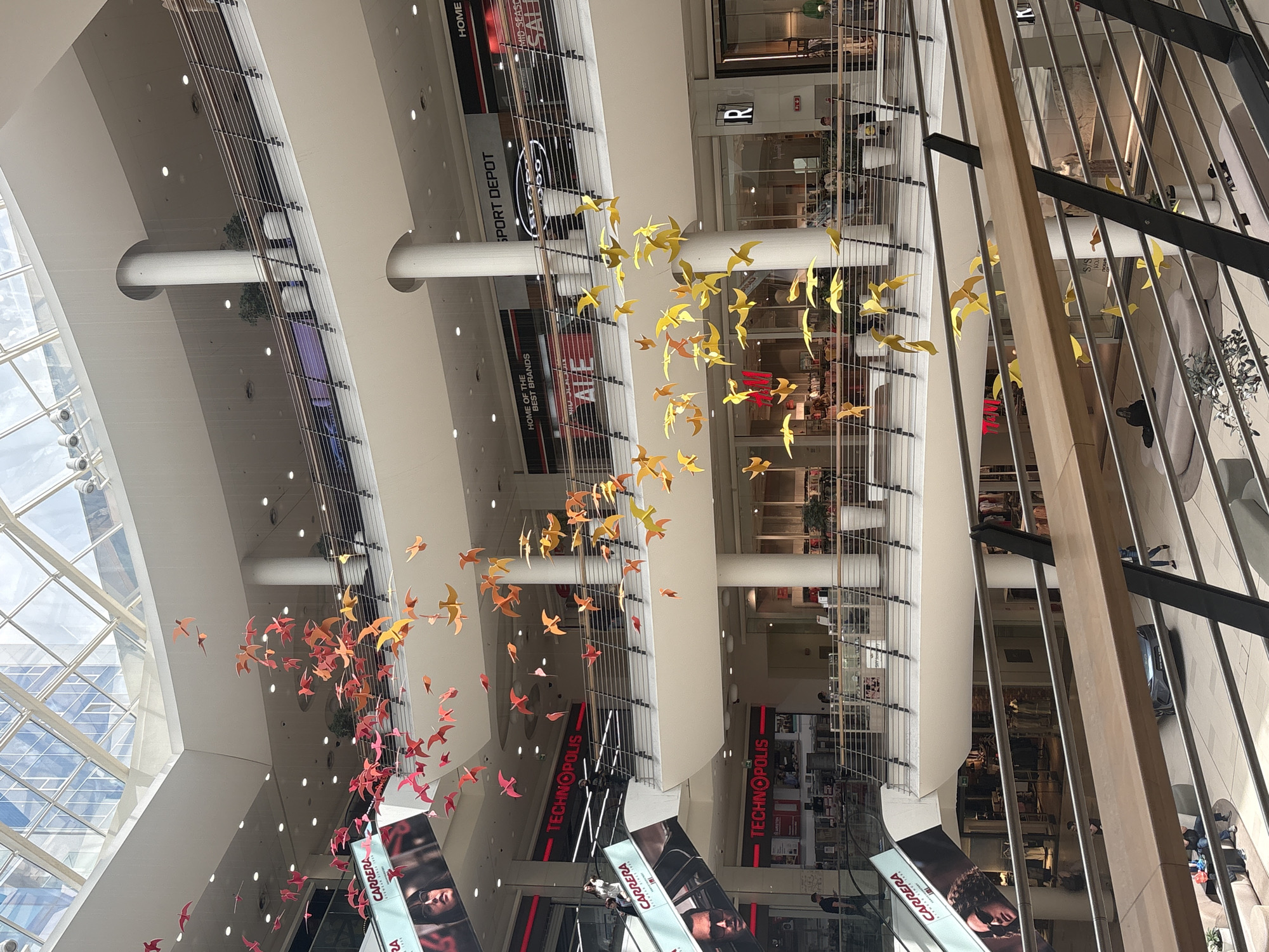 This is an interior shot of a multi-story shopping mall, with a focus on the decorative art installation of hanging, colorful bird figures. The installation features birds in shades of red, orange, and yellow, suspended from the ceiling, creating a sense of movement. The mall itself has multiple levels, visible through the metal railings and support beams. Each level houses various retail stores, indicated by logos and signage, including "SPORT DEPOT" and "TECHNOPOLIS", and advertising banners with "CARRERA" visible. Overhead, a skylight provides natural light. People can be seen on different levels of the mall, moving about. The overall architectural style of the mall is modern, with clean lines and neutral color tones.