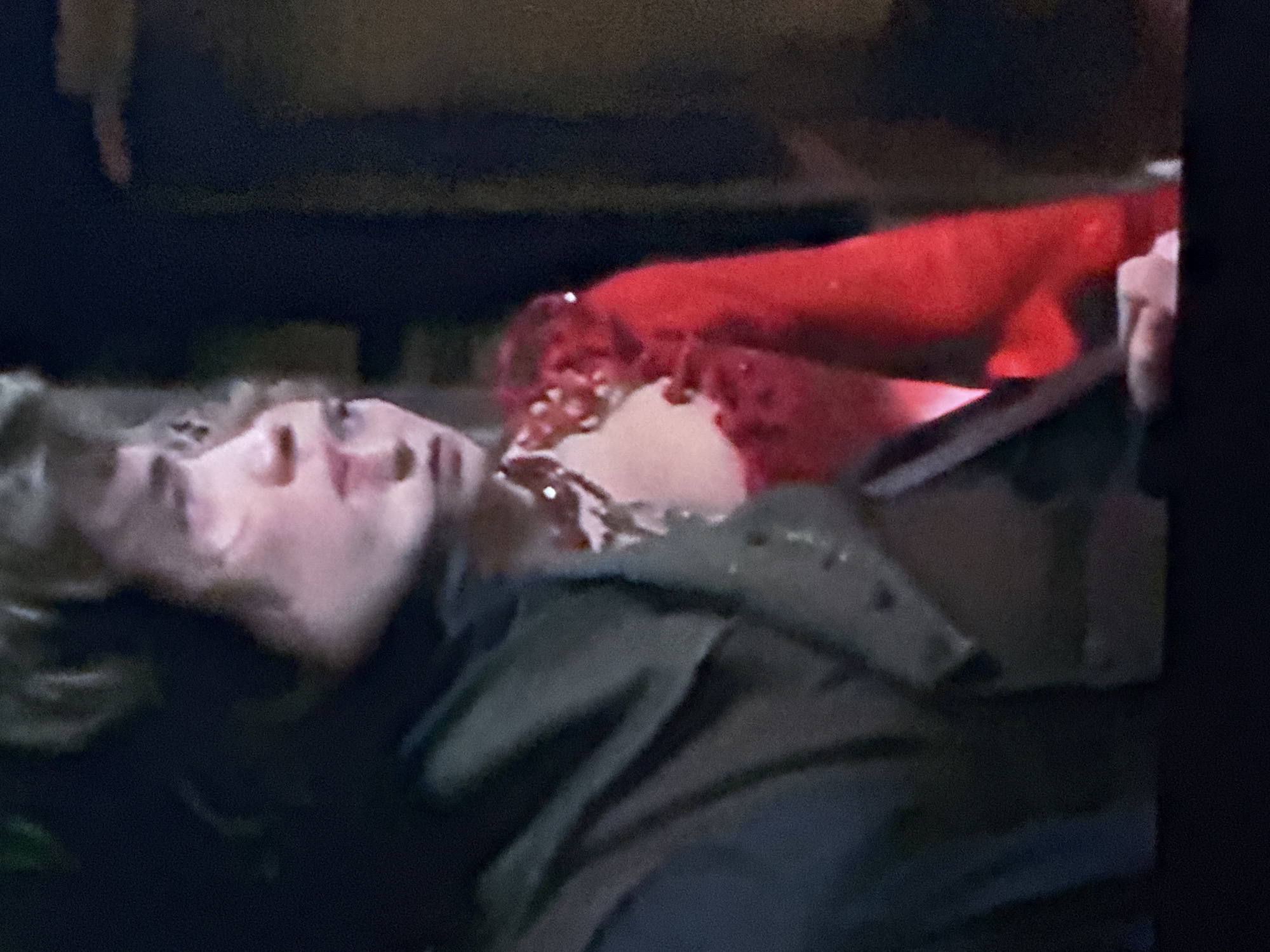 A man lies supine, his head tilted back, with apparent blood or red liquid around his neck and chest. He is wearing a dark-colored jacket or coat. A hand holding an object, potentially a metal or dark-colored tool, is visible on the right side of the image. The background is dark and indistinct. No specific details within the image indicate a location in Ivanteyevka, Russia.