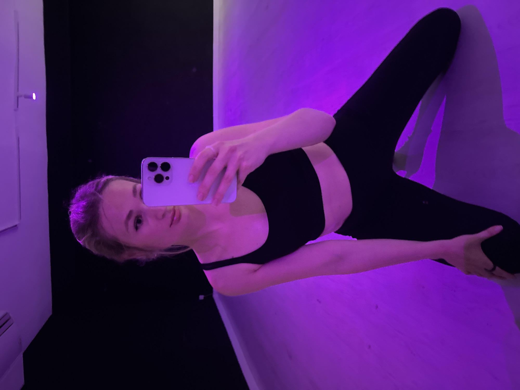 A young woman with blonde hair pulled back in a bun takes a mirror selfie in a dance studio. She is wearing a black sports bra and black leggings, and is kneeling on a wooden floor that is illuminated by purple lighting. She is holding a light purple smartphone in her right hand to take the photo. The background is dark, with a black wall and ceiling, and some track lighting visible. The scene is likely inside a dance studio or gym.