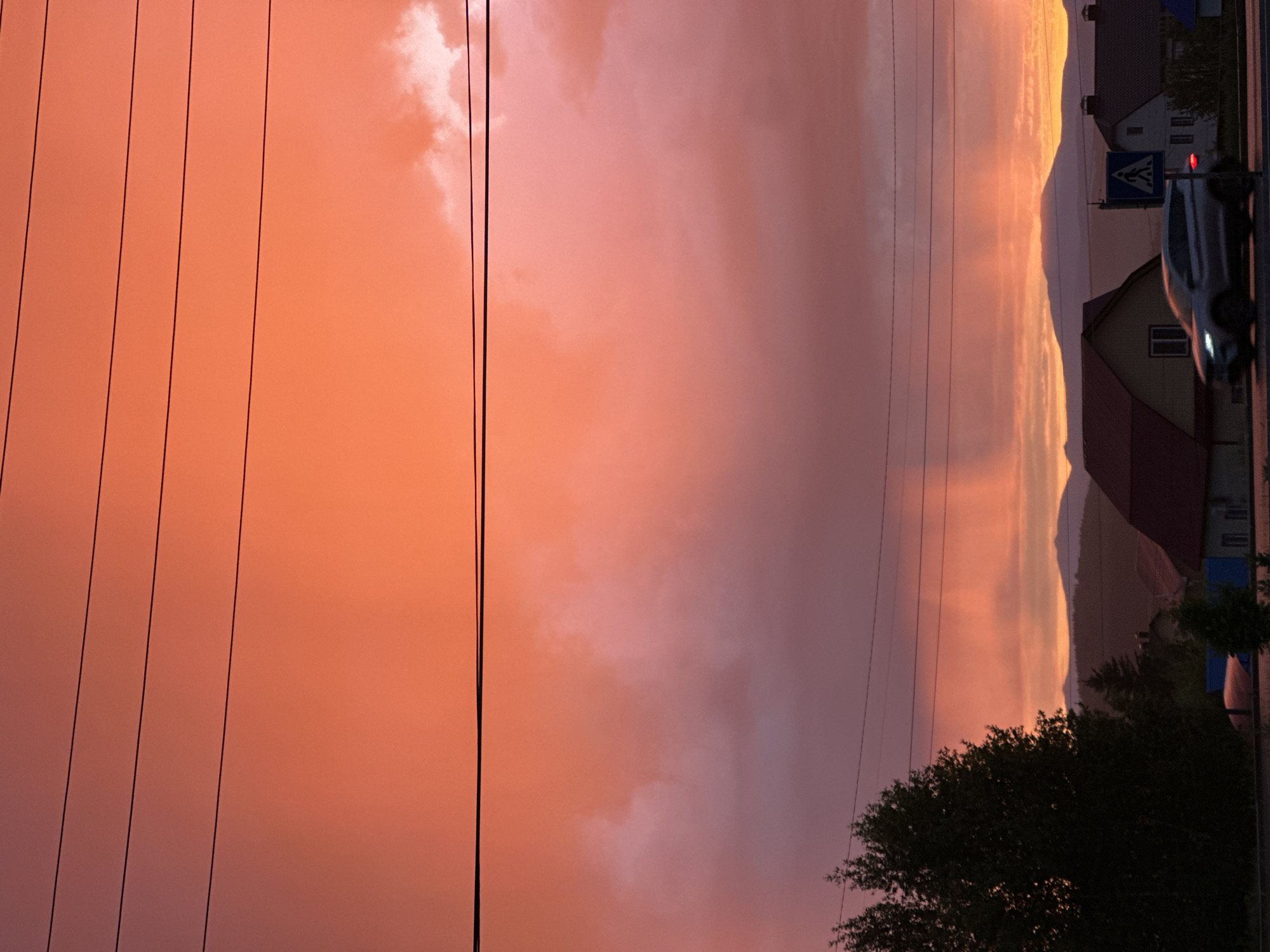 The image captures a vibrant sunset or sunrise over a semi-rural or suburban landscape, dominated by a vividly colored sky. The sky transitions from bright orange and yellow tones on the left to softer pinks and muted purples towards the right, with scattered clouds reflecting the warm light. Numerous dark power lines stretch horizontally across the upper and middle portions of the sky, creating distinct parallel lines against the colorful backdrop.
In the lower right section of the image, a scene unfolds beneath the dramatic sky. A silhouette of distant mountains or hills is visible along the horizon, their peaks softened by the atmospheric conditions. Below the mountains, several houses or buildings with varying rooflines are partially visible, suggesting a residential area. A paved road runs along the bottom edge, where a light-colored car, possibly white or silver, is in motion with its headlights illuminated. Adjacent to the road, a blue square sign featuring a white pedestrian crossing symbol is clearly visible. Dark green foliage, including a prominent tree in the bottom left corner and smaller bushes, dots the landscape, adding contrast to the bright sky. The presence of the car with its headlights on, coupled with the intense sky colors, strongly indicates either dawn or dusk. No discernible text is visible other than the pedestrian crossing symbol.