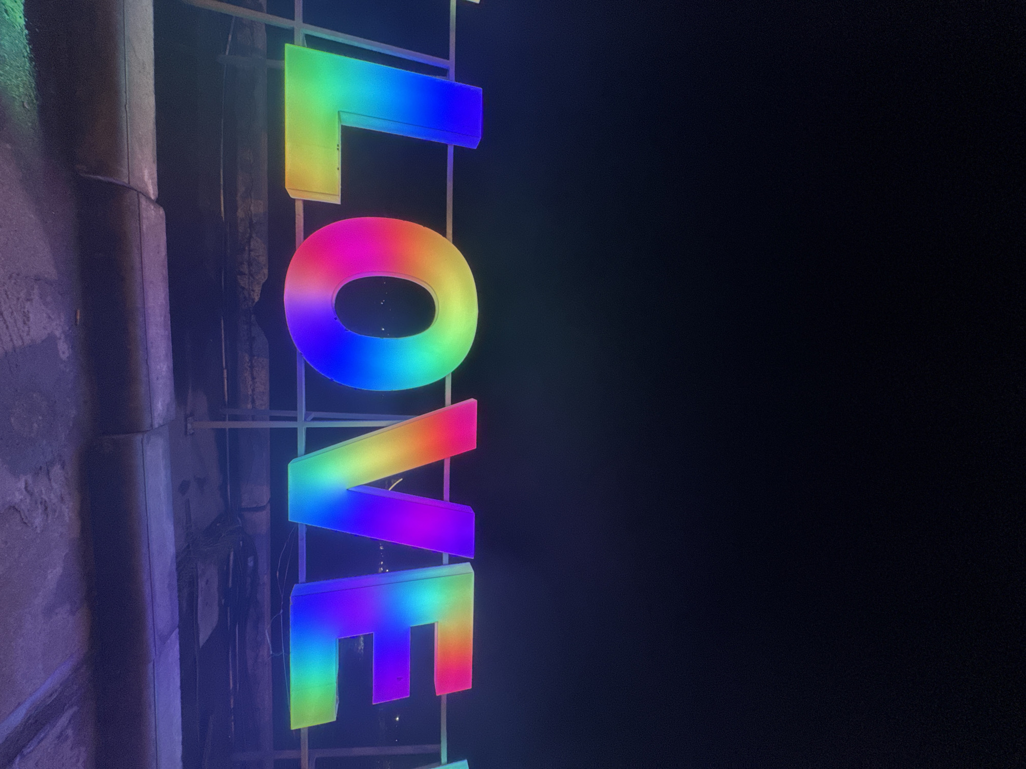 The image displays an outdoor scene at night in ตำบลเกาะพะงัน, Thailand, featuring a large, illuminated sign spelling "LOVE". Each three-dimensional capital letter is lit with a gradient of rainbow colors. The 'L' transitions from blue and indigo at the top to green and yellow at the bottom. The circular 'O' exhibits a full spectrum, including reds, oranges, yellows, greens, blues, and purples. The 'V' and 'E' letters display similar rainbow gradients, with vibrant pinks and reds at their upper left portions fading into oranges, yellows, greens, and blues towards their lower and right sections.
The letters are mounted on a light-colored, metallic railing or support structure. Behind the letters, several dark wires are visible, indicating their electrical connection. The background consists of a uniformly dark, near-black sky, occupying the upper two-thirds of the frame. A subtle, very dark blue horizontal line is discernible behind the letters, suggesting a distant horizon, with faint, scattered pinpricks of light visible below it. The ground directly beneath the sign is a dark, textured surface, possibly concrete, exhibiting faint purple and blue reflections from the sign's illumination.