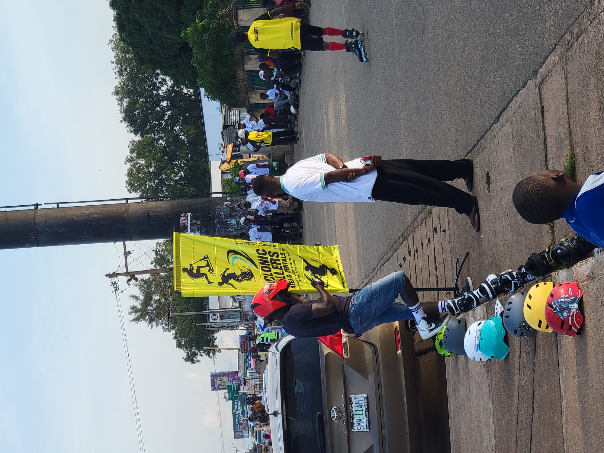 This image depicts a street scene in Jos, Nigeria, likely near a busy intersection or thoroughfare, given the presence of vehicles, advertising billboards in the background, and a significant gathering of people. A prominent yellow banner, featuring graphics of individuals on scooters and unicycles, advertises "CYCLONIC ROLLERS" and "SKATES & RENTALS," suggesting an event or business related to inline skating or similar activities. Several people are present, some wearing rollerblades, others watching. A man in a red helmet stands near a brown Toyota Venza car, appearing to interact with someone holding a phone. Lined up on the sidewalk are numerous helmets of various colors, some with designs, and a set of rollerblades. The overall atmosphere appears to be a casual outdoor gathering, possibly a community event or promotional activity.