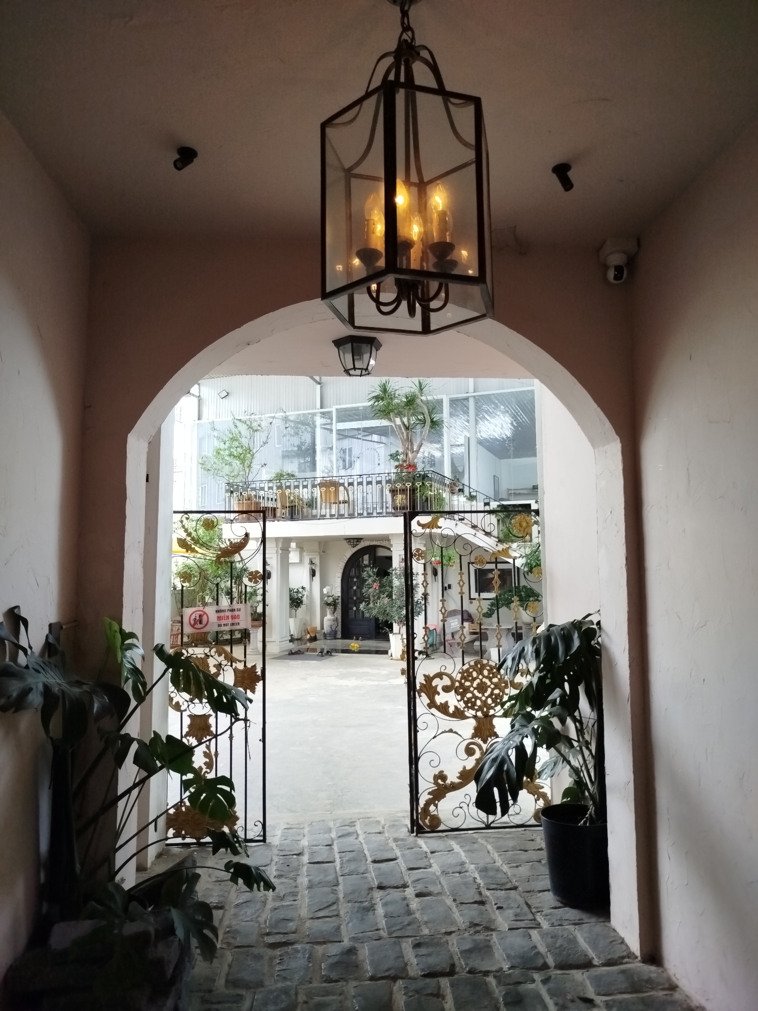 The image depicts an arched entryway to an outdoor courtyard at what appears to be a restaurant or cafe in Phường Xuân Hương, Đà Lạt, Vietnam. The entryway is paved with stone bricks. A large, dark metal lantern with four illuminated bulbs hangs from the ceiling. On the left side of the entryway, a large potted plant with broad green leaves is visible. On the right side, a smaller potted plant is also present.
The courtyard is accessible through an ornate black metal gate, which is partially open. The gate features intricate scrollwork with gold accents. A sign on the gate reads "KHÔNG PHẢN SỰ MIỀN VÀO" and "DO NOT ENTER" in English, indicating restricted access.
Beyond the gate, the courtyard features a paved area with some scattered shoes. In the background, a building with large glass windows and balconies is visible, with several trees and potted plants adorning the space. A security camera is mounted on the wall to the right of the entryway, and another smaller camera is visible on the ceiling above the arch. The overall impression is a well-maintained, inviting, and somewhat upscale establishment.
