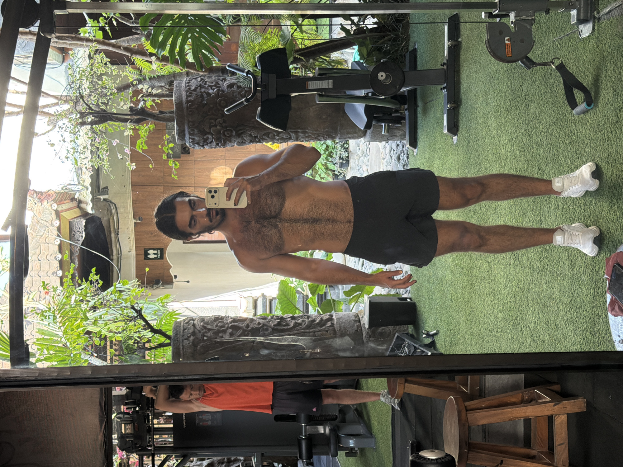 A man is taking a mirror selfie in an outdoor gym. He is wearing black shorts and no shirt. In the reflection, another man is visible on a weight machine, wearing an orange tank top and black shorts. The gym has green turf flooring and is surrounded by lush vegetation and stone pillars. The image captures a moment of fitness in a tropical setting within North Kuta, Indonesia.