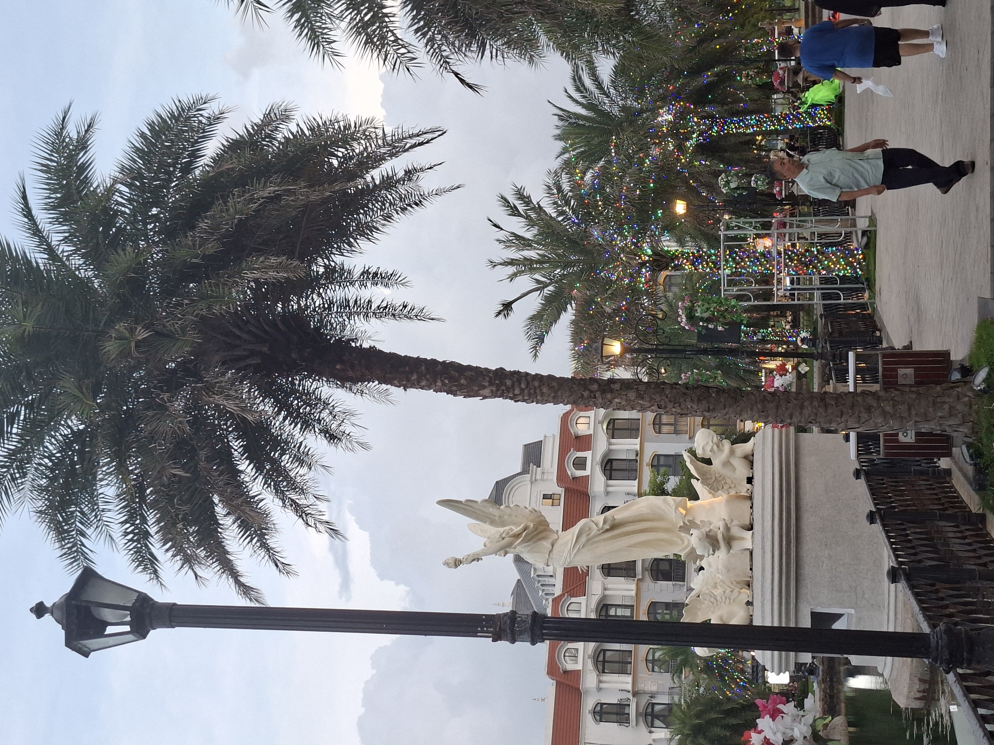 The image shows an outdoor scene in the city of Phú Quốc, Vietnam. In the foreground, a lamppost stands to the left, next to a statue of a winged figure holding a torch, supported by smaller winged figures. The statue is positioned near a body of water and a dark, ornate fence. A palm tree with dense fronds dominates the right side of the image. Behind the statue and palm tree, a multi-story building with red-tiled roofs and white facades is visible. The background features several palm trees decorated with numerous colorful lights, suggesting a festive atmosphere. Two men are walking on a paved path in the middle ground, one wearing a light green shirt and dark pants, the other a blue shirt and dark shorts. The sky is overcast.