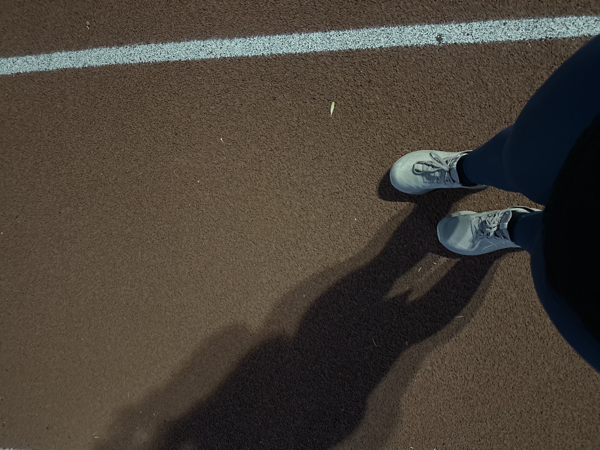 The image presents a top-down view of a person's feet and lower legs on a running track in Puebla, Mexico. The person is wearing light grey athletic sneakers with laces and dark-colored athletic pants. The running track has a distinct textured, rubberized brown surface, characteristic of an athletic facility. A clean, white lane line runs horizontally across the upper portion of the frame, marking a boundary on the track.
Long, dark, and well-defined shadows, cast by the person, stretch diagonally across the track towards the lower left, indicating a strong, low-angle light source. This suggests the time of day is either early morning or late afternoon/evening. The overall lighting appears somewhat dim, which could imply evening conditions, possibly under stadium lights that are powerful enough to create sharp shadows. The clarity of the shadows also suggests clear weather. A small, light green, leaf-like piece of debris is visible on the brown track surface, slightly below the white line and to the left of the right foot. No text is visible in the image. The person appears to be standing still on the track.