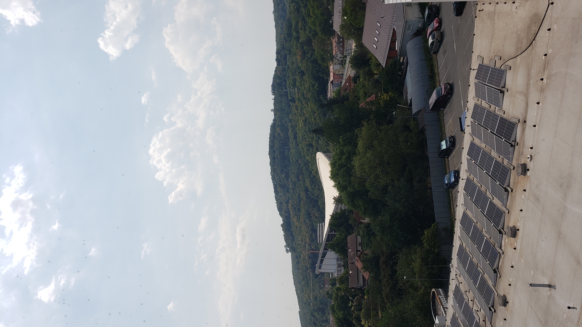 The image captures a view of the city of Prešov, Slovakia, specifically looking towards the stadium "City Stadium" (Štadión Tatran). In the foreground, a rooftop is covered with numerous solar panels, arranged in rows. A circular structure with text "LASTOST" is visible on the left side of the rooftop. Adjacent to the solar panel-covered rooftop is a parking lot with several cars parked. Beyond the parking lot, a dense green forest covers a hill, with a modern stadium featuring a distinctive white curved roof prominently situated on the hillside. Several residential buildings with tiled roofs are scattered among the trees. The sky above is partly cloudy with white cumulus clouds against a light blue backdrop.