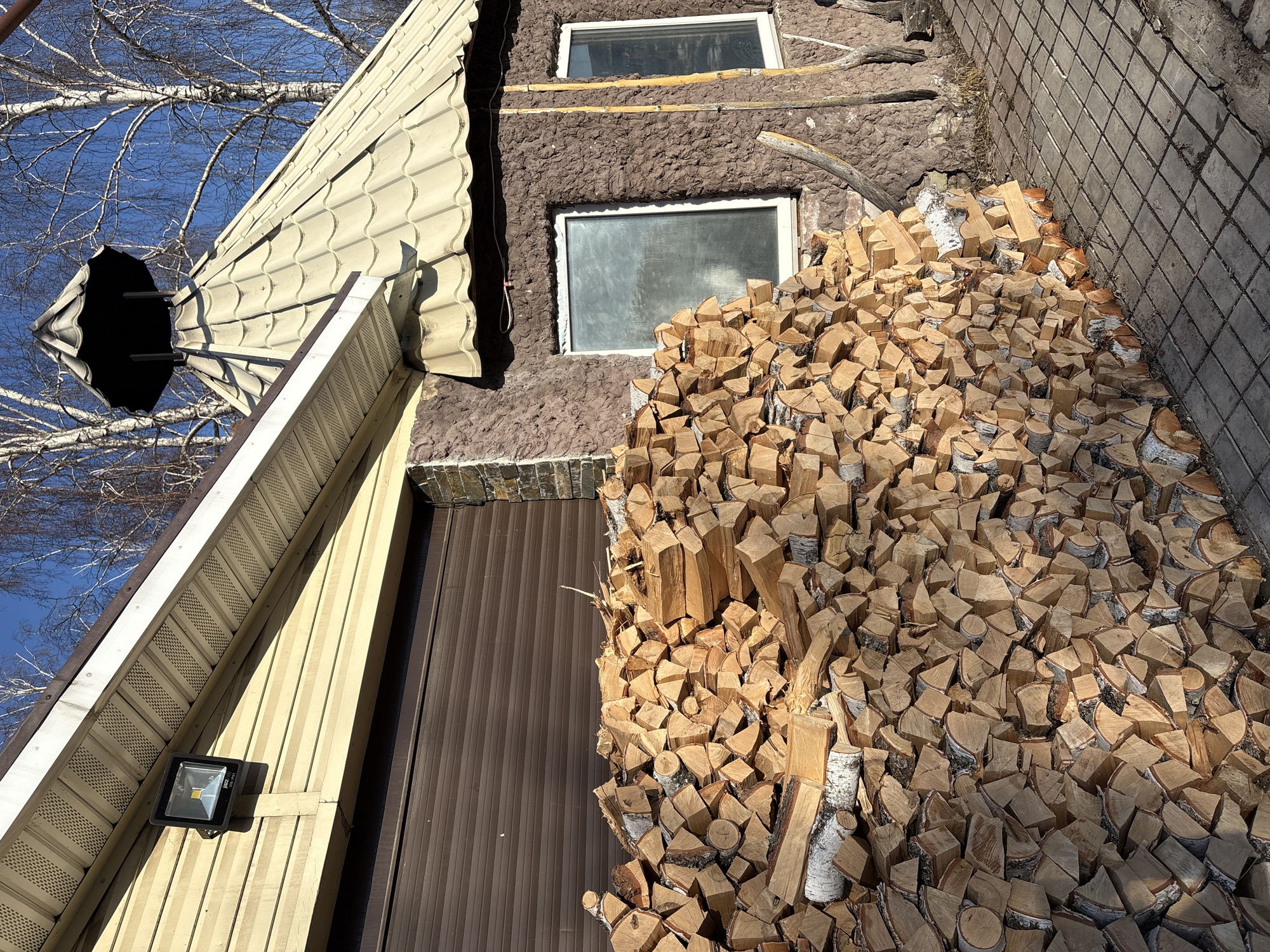 The image depicts an exterior scene in Bely Klyuch, Russia, featuring a building with a large stack of firewood under a clear blue sky.
The building's left section presents a wall clad with light beige horizontal siding, below which is a brown, horizontally corrugated roll-up door. A black rectangular LED floodlight is mounted on the beige siding, featuring visible internal LED elements. The building's corner transitions to a darker, stone-textured cladding.
The right section of the building displays a brown, heavily textured stucco-like wall. Two white-framed rectangular windows are set into this wall; the left window appears frosted or obscured, while the right window reflects the environment. A weathered wooden pole or branch leans against the wall to the right of the windows. The building is topped with a conical roof covered in light beige, wavy-patterned tiles, culminating in a dark, bell-shaped ornamental structure at its apex.
In the foreground, a substantial pile of neatly stacked, light-colored firewood, primarily split logs with visible birch bark, extends along the base of the building, partially obscuring the lower portions of the textured wall and the left window. The ground is paved with square grey concrete pavers, reflecting direct sunlight and casting shadows.
The background reveals bare deciduous trees, primarily birches, against a vibrant, cloudless blue sky, indicating a season devoid of foliage. No individuals or active subjects are present in the scene.