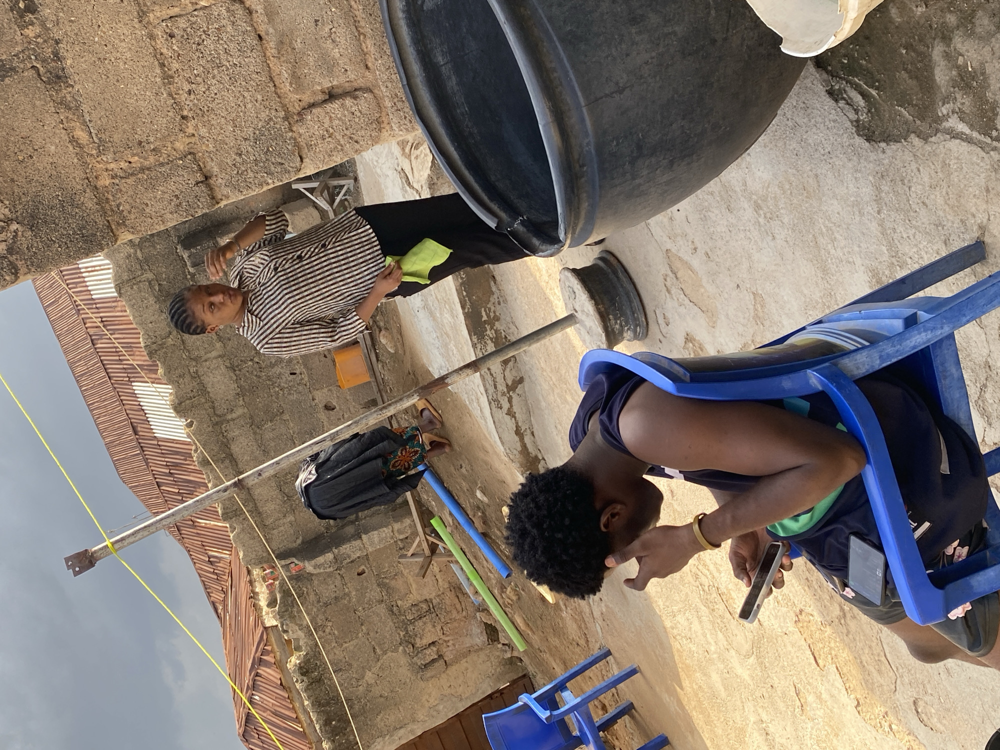 In Jos, Nigeria, a young man is seated on a blue plastic chair, engrossed in his mobile phone, with another phone resting on his lap. He is wearing a dark sleeveless shirt and patterned shorts. Beside him, a young woman in a striped blouse and black pants stands near a large black plastic basin. She holds a green cloth in her hand, and appears to be talking. The background shows a wall constructed of cement blocks, a corrugated metal roof, and a metal pole with a yellow rope strung between it and another support. Two blue and green pipes lie on the ground, and a person in a dark skirt and patterned attire is visible in the distance. The ground is made of concrete and dirt.