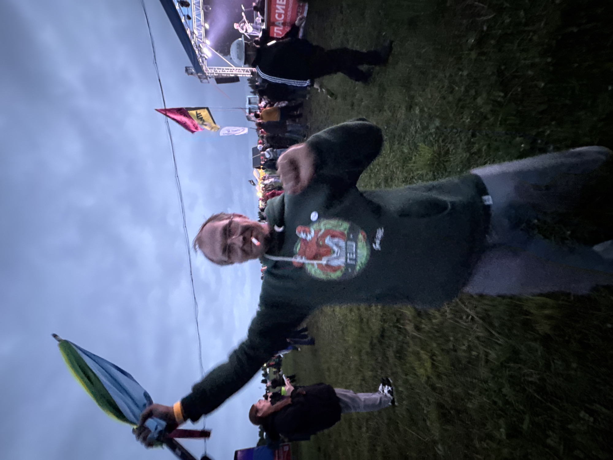 In Bol'shaya Kivara, Russia, an outdoor evening scene depicts a man in the foreground, center-left, with glasses, a dark green hooded sweatshirt featuring a red fox graphic and "FEW" text, and grey pants. He holds a partially folded umbrella, striped yellow, green, and blue, aloft in his raised right hand, displaying a yellow wristband. A lit cigarette is in his mouth as he looks toward the camera with a dynamic pose. To his left, a woman with reddish-brown hair, wearing a dark jacket and light pants, stands looking at a mobile device while holding a green cup. To the right, a man in a white helmet, a black track jacket with white arm stripes, and dark pants walks right, carrying a bottle. In the background, an illuminated stage features a band performing. Nearby, a red and yellow flag and a white flag are visible, along with a banner partially displaying the Cyrillic text "ГЛАСИЕ И ОБЩЕСТВЕННОЕ." Multiple indistinct figures populate the vast grassy field under an overcast sky, suggesting a public event or concert. An electrical wire stretches across the upper sky.