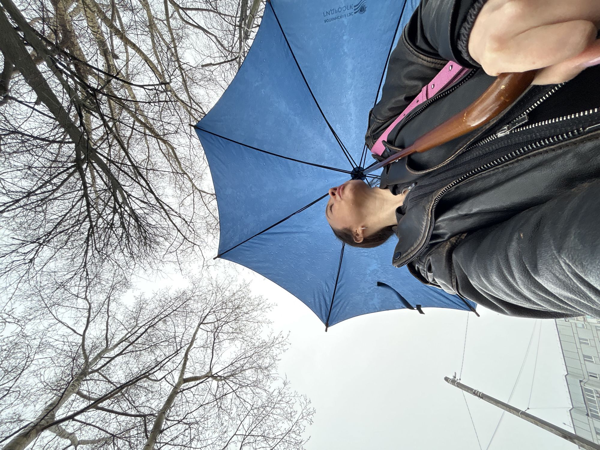 A low-angle shot depicts a woman holding a blue umbrella on a cloudy day. The woman wears a black leather jacket with a pink strap visible beneath. Bare tree branches fill the left side of the frame against a gray sky. In the lower right corner, a utility pole and a gray building with white-framed windows suggest an urban environment. The woman's face is visible under the umbrella as she looks upwards.
Based on information from the text on the umbrella, the location is Chaikovsky, Russia.