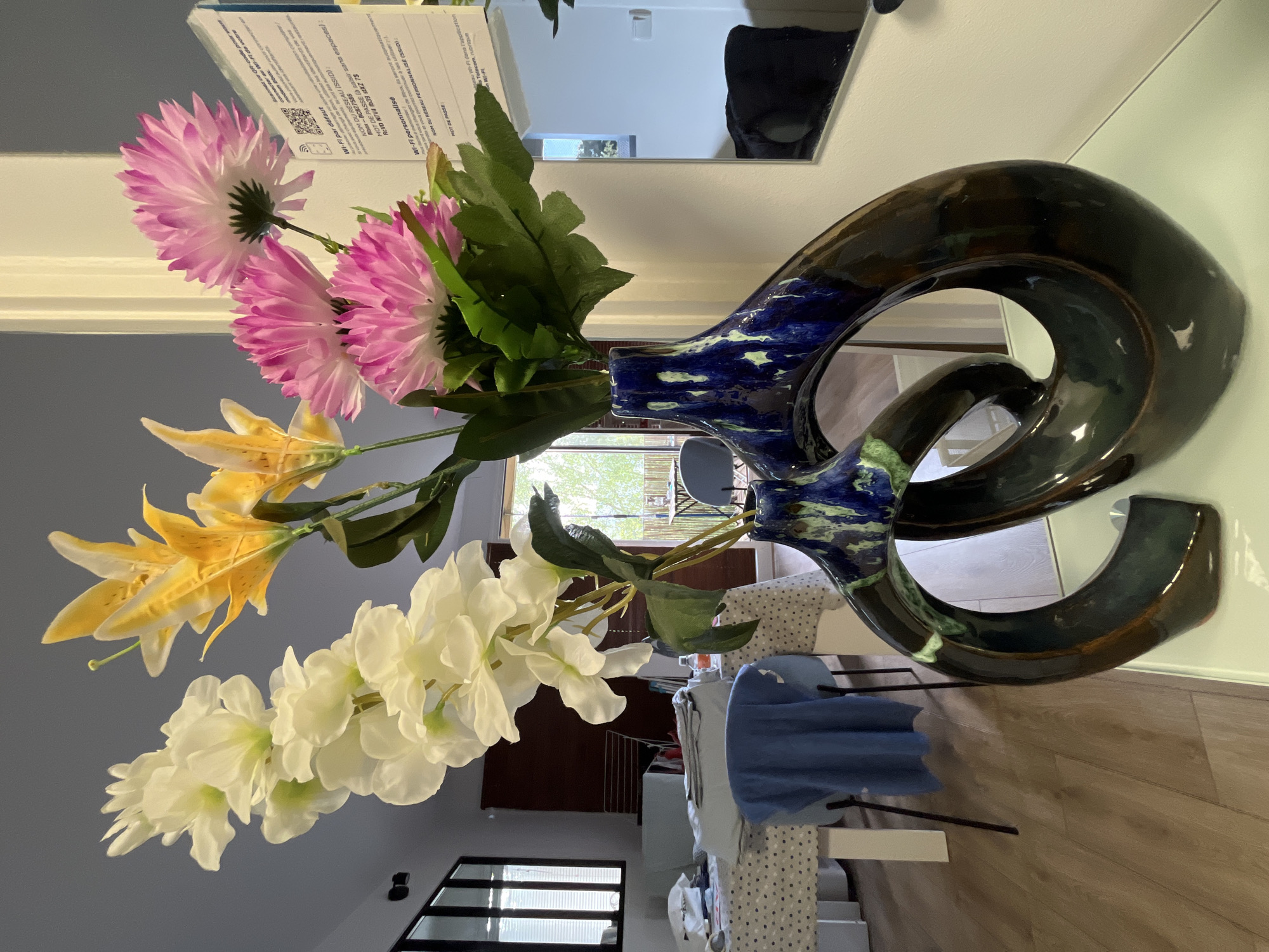 This is a vertically oriented, eye-level, medium shot of an artificial flower arrangement in a decorative vase. The vase is a glossy, dark brown, and features a unique, twisted, abstract shape with a blue and green marbled glaze on its upper portion. The flowers are arranged in bunches, with a cluster of bright pink and white flowers at the top, followed by yellow lilies, and a large spray of white flowers at the bottom. The background shows a domestic interior, with a wall, a mirror reflecting a room, and a table with a patterned tablecloth. There is some text visible in the upper left corner of the image, appearing to be instructions for Wi-Fi connection, including a QR code. The lighting appears to be natural, suggesting daytime. The overall impression is one of a still life arrangement within a home setting.