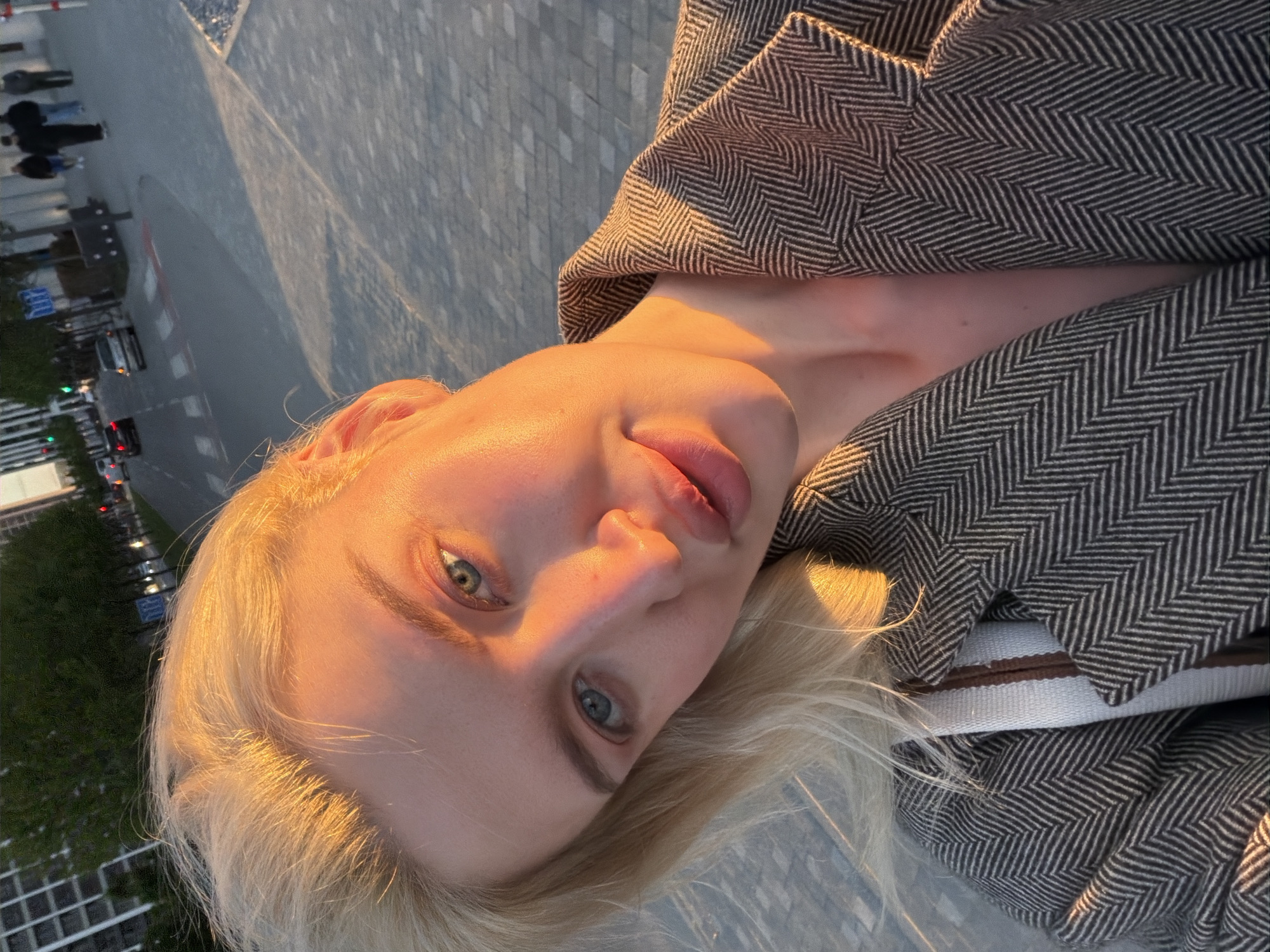 A tilted, close-up portrait captures a young woman with blonde hair and captivating blue-green eyes, looking directly at the viewer. Her fair skin is bathed in the warm, golden light of what appears to be the golden hour, creating soft highlights on her forehead, nose, and cheekbones, and casting gentle shadows. Her lips are slightly parted.
She is dressed in a structured coat or jacket featuring a prominent grey and brown herringbone pattern. A white strap with a central brown stripe, possibly from a bag, is visible over her shoulder. The camera angle is high, looking down at her, and the entire image is rotated, suggesting a dynamic selfie or a carefully composed shot.
The background is an out-of-focus urban environment, depicting a paved area that could be a sidewalk or plaza, with a street and faint outlines of vehicles and other pedestrians in the distance. Tall buildings are barely visible on the far left. The soft, warm light and the urban scenery suggest the image was taken in a city like Belgrade, Serbia, during either sunrise or sunset. There is no visible text in the image.
