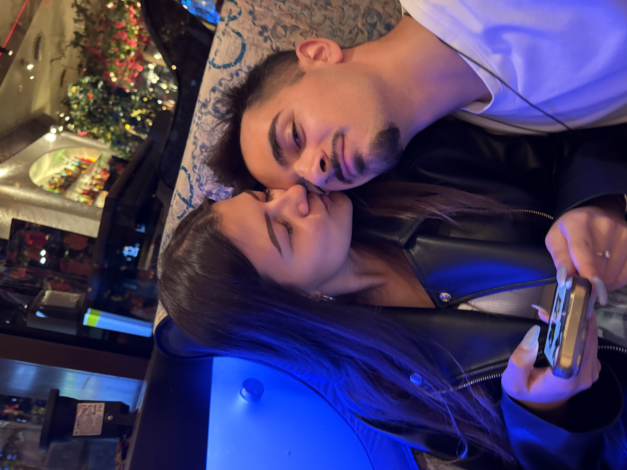 A young couple is captured in a close-up, dimly lit scene within what appears to be a modern restaurant or bar in Sofia, Bulgaria, likely a Happy Bar & Grill establishment given the distinct decor. The woman, positioned on the left, has long dark hair and is wearing a black leather jacket over a white top. She is gently kissing the man on his right cheek, her eyes closed in affection. Her left hand, adorned with long, light-colored acrylic nails and a ring, holds a small, gold-colored electronic device. The man, on the right, looks directly towards the camera with a subtle smile, sporting short dark hair, a mustache, and a chin beard. He wears a white t-shirt and a thin dark chain necklace. He leans his head back against a light-colored, patterned cushion. The background on the left features a prominent, curved panel emitting a bright blue light, with a dark, cylindrical object mounted above it. To the right, beyond the couple, the blurred background reveals an arched interior design, shelves filled with numerous colorful bottles, and decorative green and red foliage adorned with small lights, indicative of a bar area.