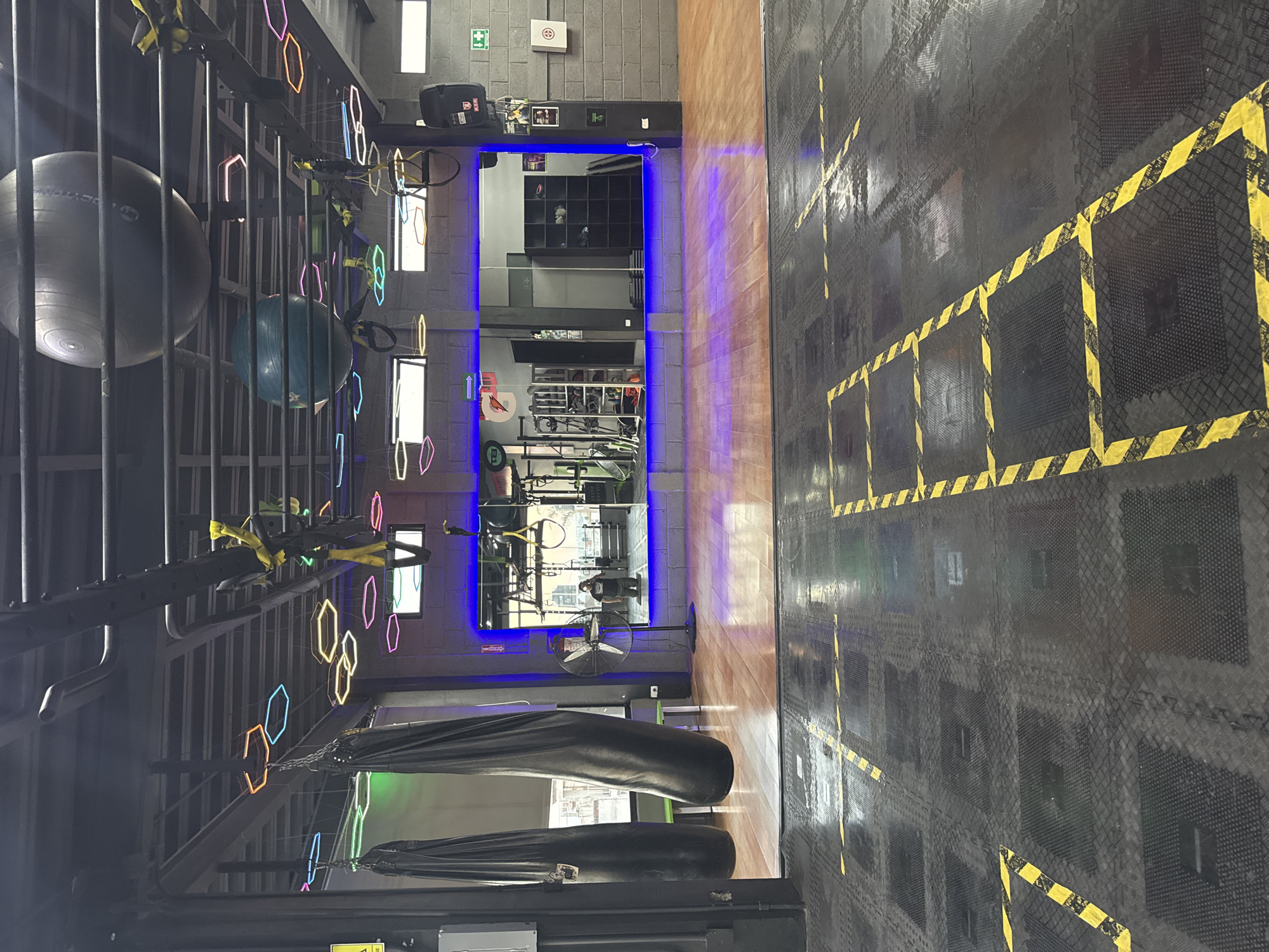 The image depicts the interior of a fitness center in Puebla, Mexico. The space contains workout equipment, including stability balls, suspension training systems, punching bags, and weight training machines reflected in a wall mirror. The floor is composed of black rubber tiles with yellow hazard markings. The facility has neon lighting along a metal frame structure. A fan is visible near the mirror. A person can be seen in the mirror.