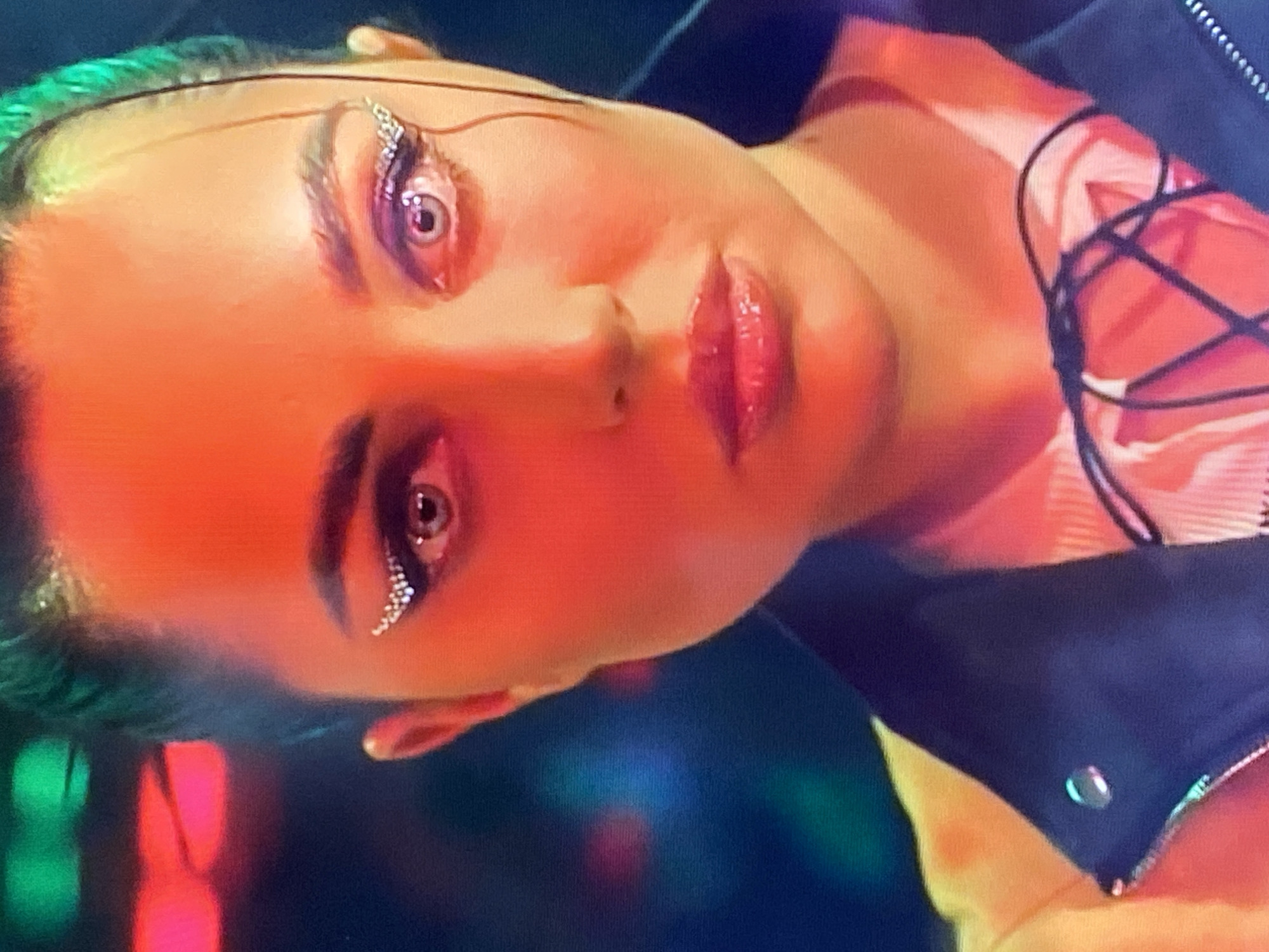 The image shows a close-up of a woman's face, illuminated by bright, colorful lights that create a neon-like effect. She has striking makeup, including rhinestones applied to her eyelids in a wing-like design. Her lips are glossy and slightly parted. She is wearing a dark jacket over a light-colored top with a criss-cross lacing detail. The background is blurred, with streaks of green and pink light suggesting an urban environment, possibly a club or entertainment venue. There is no specific information in the image to indicate the location is Yekaterinburg, Russia.
