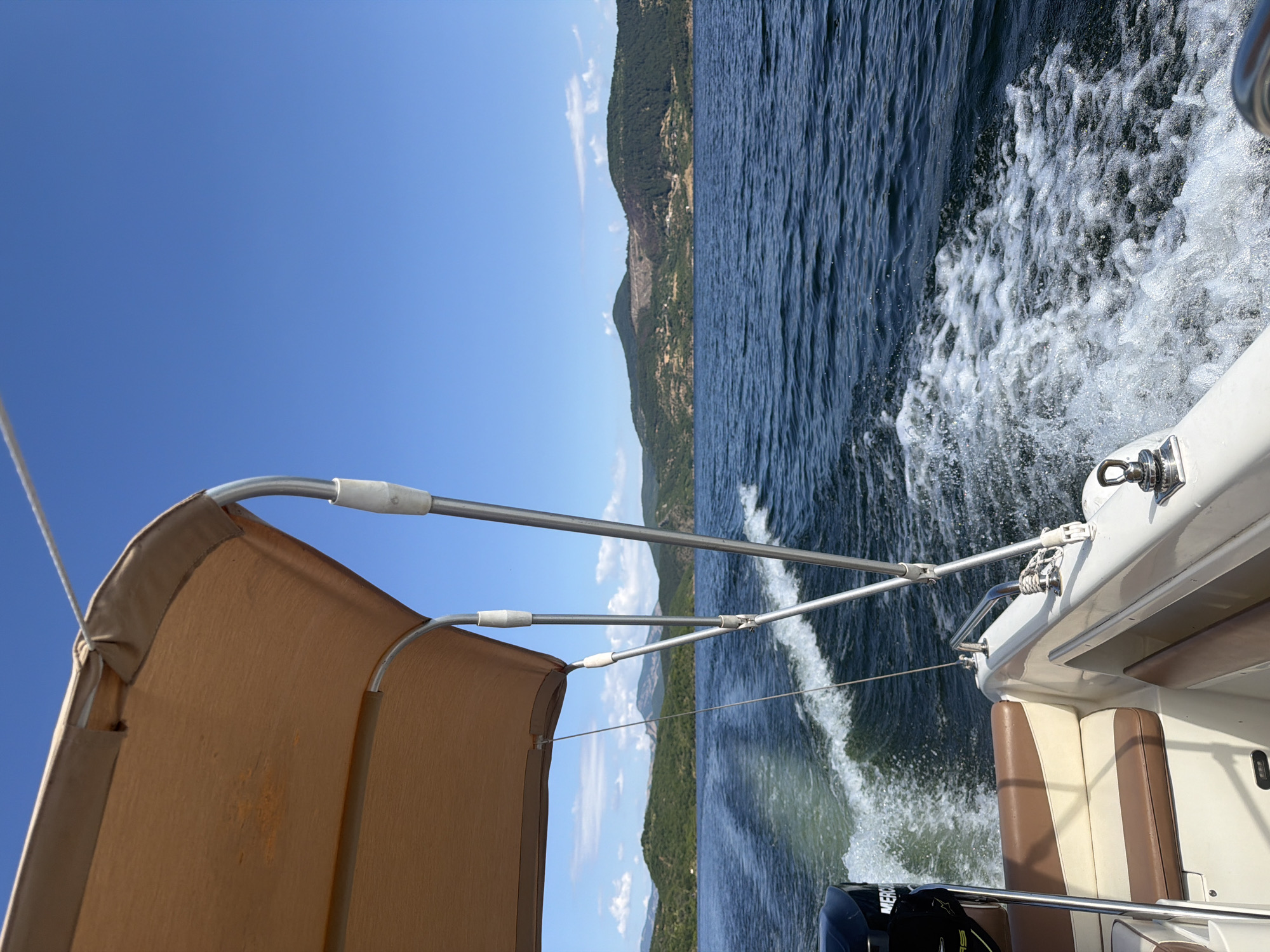The image shows a view from a boat moving through a body of water. The boat has a tan canopy supported by silver poles. The seats of the boat are visible and appear to be upholstered in white and brown. The wake of the boat is prominent in the water, creating a trail of white foam. In the background, there are green, tree-covered hills under a bright blue sky with some white clouds. The overall scene depicts a sunny day on the water, likely at the Yovkovtsi Reservoir near Glavatartsi, Bulgaria. The outboard motor of the boat, a Mercury, is partially visible on the left side of the image.