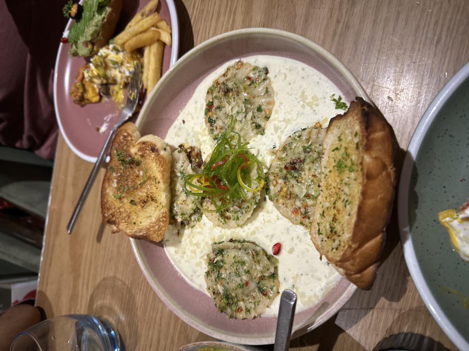 The image displays an indoor restaurant dining scene on a light-colored wooden table. A large, round, pink-rimmed plate centrally features a dish with a creamy white sauce. Within the sauce are five ravioli-like pieces, garnished with chopped green herbs, red chili flakes, and grated white cheese. The central piece includes additional julienned green and yellow strips with red threads. Two toasted garlic bread slices, topped with green herbs, are positioned at the plate's top and bottom. A single red pomegranate seed rests in the sauce.
To the upper right, a smaller, dark pink plate holds partially consumed items: golden-brown french fries, a portion of a yellow and white scramble, and a green avocado toast with a black object and red seed garnish. A silver dinner fork is placed on this plate. A clear, blue-tinted glass of water is visible to the upper left. The handle of a silver utensil is on the main plate's left edge. The rim of a light green, speckled bowl with yellow and white food is partially visible at the bottom.
Blurred figures, one in a reddish-brown garment, are present in the background. The specific city of Kalyan-Dombivli, India, cannot be visually confirmed from the image content.