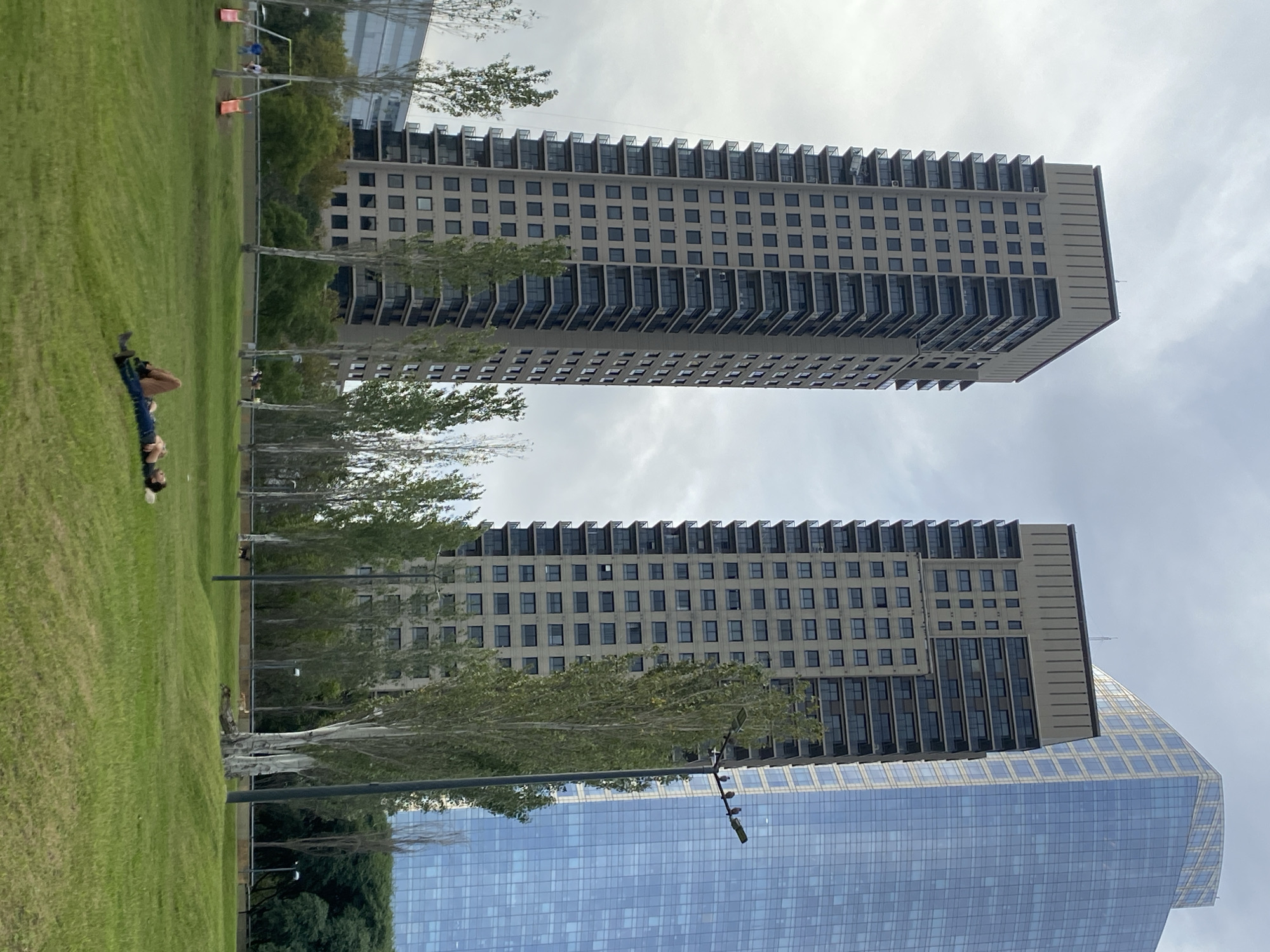 The image depicts a park in Buenos Aires, Argentina, with two tall apartment buildings in the background. A person is lying on the grass. A third building, covered in glass, is also visible. There are trees in the park and streetlights. The sky is overcast.