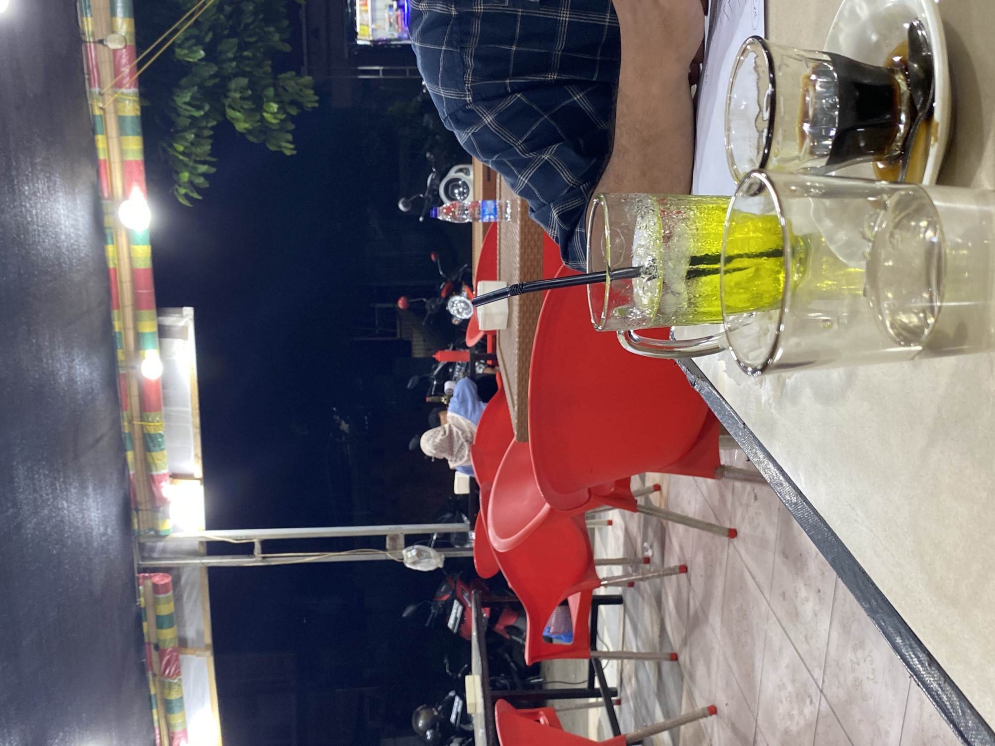 A close-up shot captures a table at an outdoor eatery in Lhokseumawe, Indonesia, at night. Two glasses are prominently featured: one containing a bright yellow beverage with ice and a straw, and another smaller glass holding a dark liquid, likely coffee, with a spoon. In the background, red plastic chairs are arranged around tables, and several motorcycles are parked. A person, partially visible from the shoulder down, wears a dark blue plaid shirt and has their arm resting on the table. The lighting suggests a casual, nighttime setting, possibly a roadside restaurant or warung. The environment appears to be open-air, with a roof structure overhead adorned with colorful decorations and string lights.