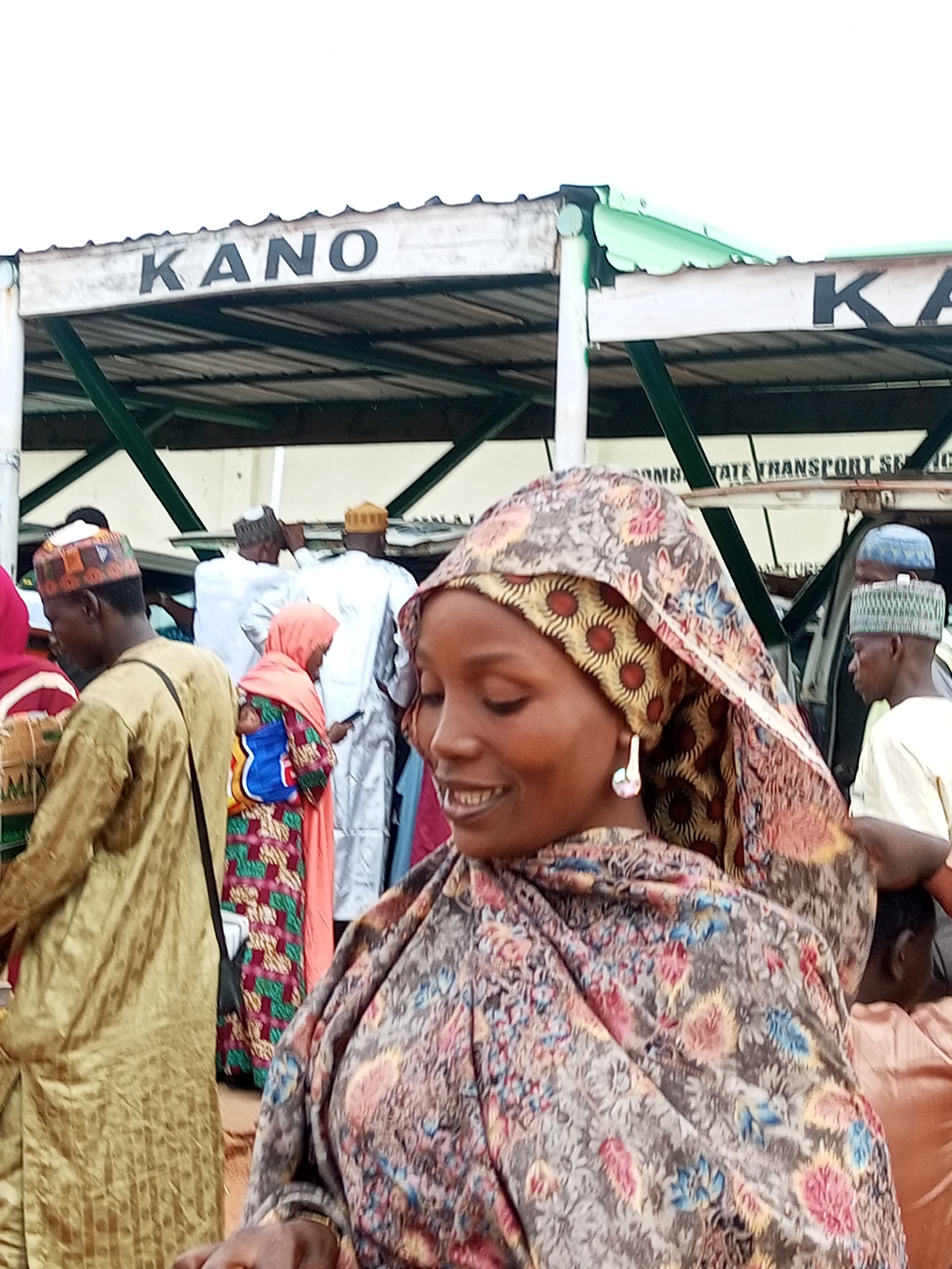 The image depicts a lively outdoor scene, likely a transportation hub or market area in Gombe, Nigeria. In the foreground, a woman with a warm smile is the central focus. She is wearing a vibrant headscarf with a floral and circular pattern in shades of pink, yellow, and brown, and a matching outfit. A prominent, teardrop-shaped earring dangles from her left ear. Behind her, a crowd of people are visible, many of them men dressed in traditional attire, including long, flowing robes and caps.
To the left, a man wearing a golden kaftan and a patterned cap is partially visible. Further back, other individuals are engaged in various activities, with some appearing to be in conversation. A sign overhead reads "KANO," possibly indicating a destination or a name associated with the location. Another partially visible sign includes text that appears to be "STATE TRANSPORT SER." The environment suggests a bustling public space, possibly a bus station or a busy market area, with structures providing shelter overhead. The overall atmosphere is one of activity and community engagement.