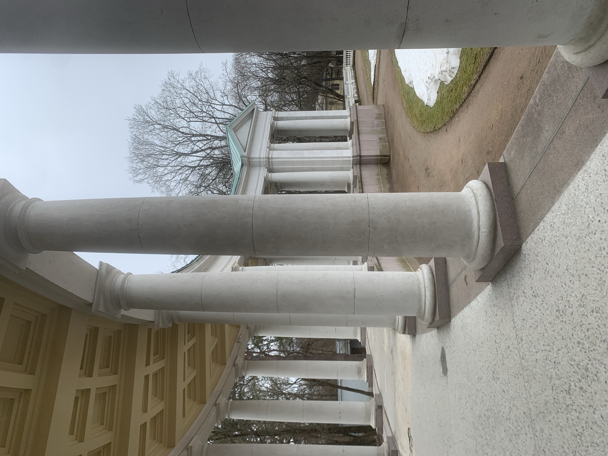 This image depicts a colonnade in Arkhangelskoye, Russia. The view is from within the structure, looking outward through a series of white columns. Beyond the columns, bare trees are visible against an overcast sky. To the right, there is a path with snow and green grass. The architecture is classical in style.