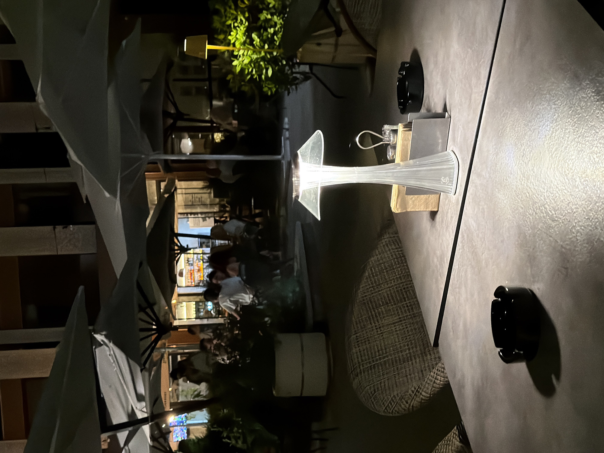 An outdoor seating area at night, possibly a restaurant or cafe, in Burgas, Bulgaria. The scene is lit by ambient lighting and a modern, translucent table lamp. In the foreground, a dark gray table has a black ashtray and the aforementioned table lamp, along with a small menu holder and what appears to be napkins. A woven chair is partially visible on the left side of the frame. In the midground and background, several tables with umbrellas are set up, and people are seated and moving around. The overall atmosphere suggests a relaxed evening setting.