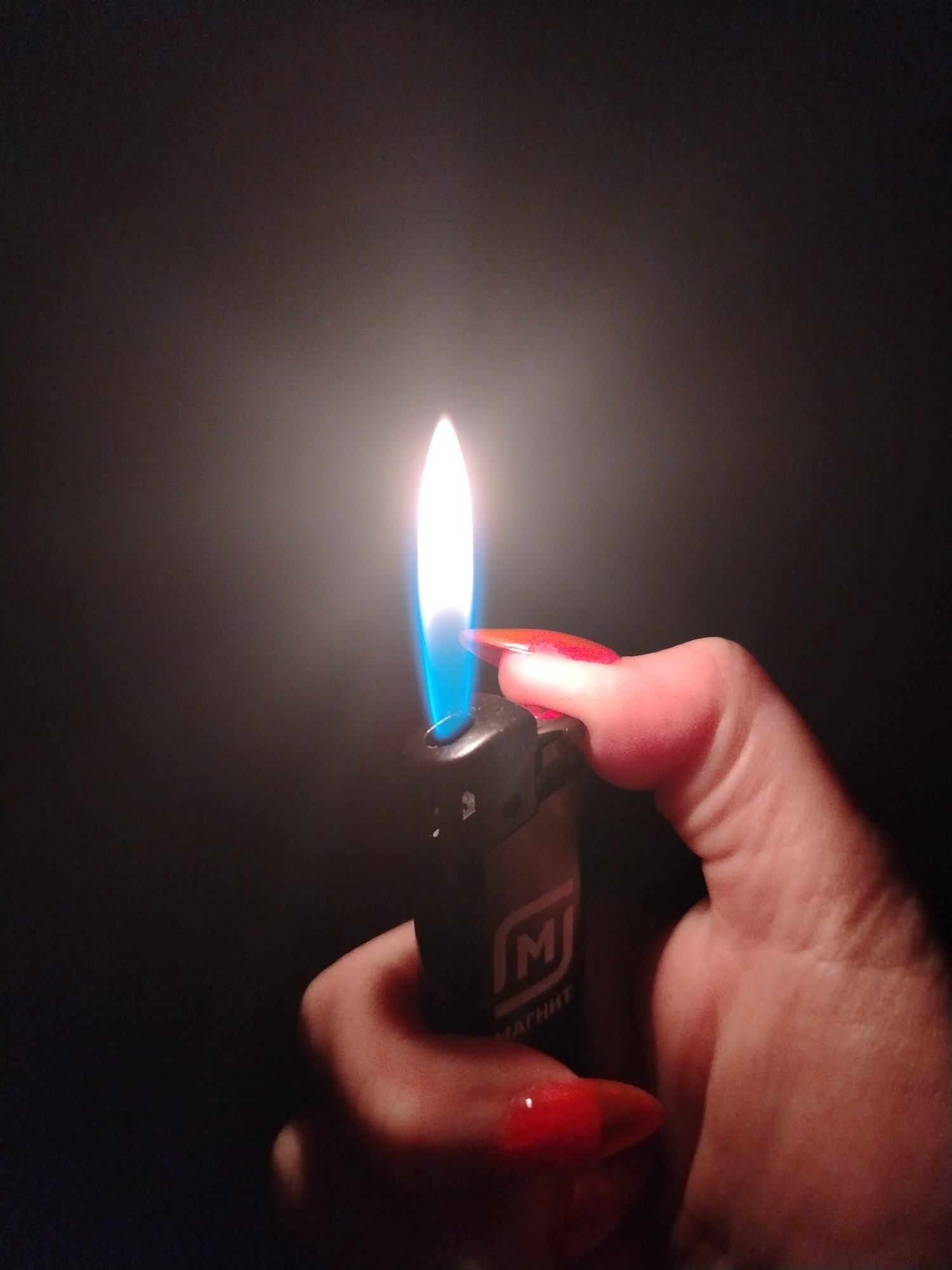 A hand holding a lit lighter is depicted against a dark background. The lighter is black with a white logo that has the letter "M" and the word "МАГНИТ" written underneath. The hand holding the lighter has red, pointed fingernails. The flame of the lighter is white with a blue base. The background is dark and featureless. There is no indication of the location being in the city of Izhevsk, Russia.