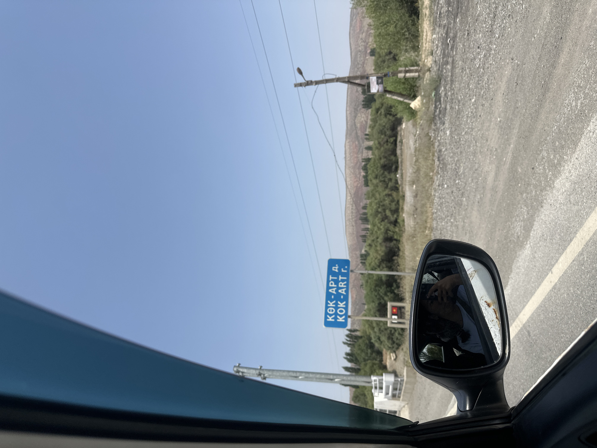 The image shows a view from inside a vehicle, looking out the side window and into the side mirror. In the side mirror, a person is reflected, taking a selfie with their phone. Outside the car, a blue road sign with white text is visible, indicating "КӨК-АРТ д. КОК-АРТ г." This sign is likely located in the city of Jany-Dyikan, Kyrgyzstan, as it appears to be a directional sign. The landscape beyond the sign consists of dry, hilly terrain with sparse trees and vegetation. Electrical poles and power lines are also present in the scene. The sky is clear and blue, indicating a sunny day. The road in the foreground appears to be unpaved, made of gravel and dirt.