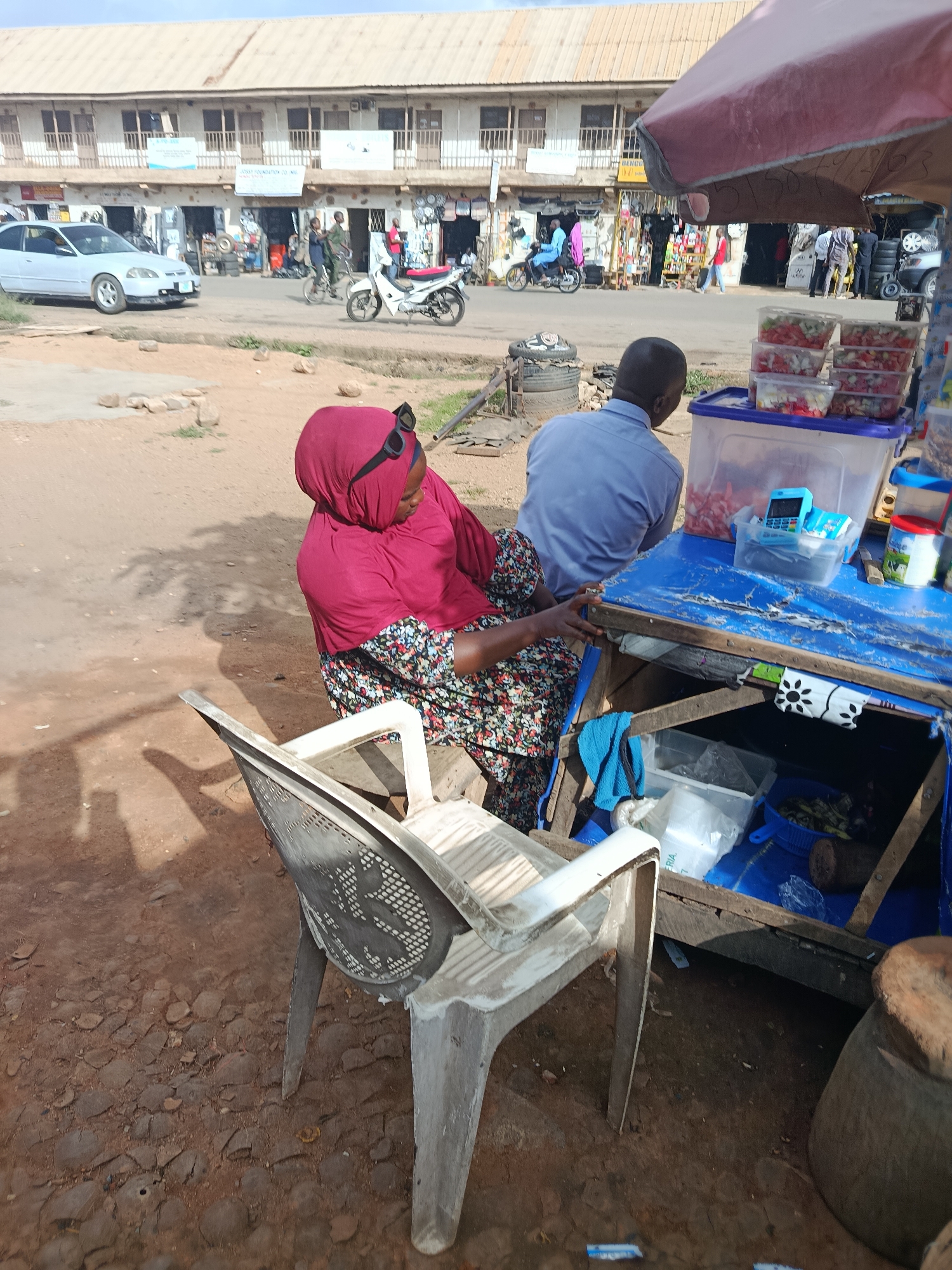 In Jos, Nigeria, a woman wearing a red hijab and a floral dress is seated on a plastic chair next to a blue market stall. She is looking down, and sunglasses are perched on top of her head. Beside her, a man in a light blue shirt is seated at the stall. The stall is laden with containers of what appear to be chopped fruits and vegetables, a blue cash register, and other items. Behind the stall, a street scene unfolds with buildings, vehicles, and other people visible in the background. The ground is a mix of dirt and scattered stones.