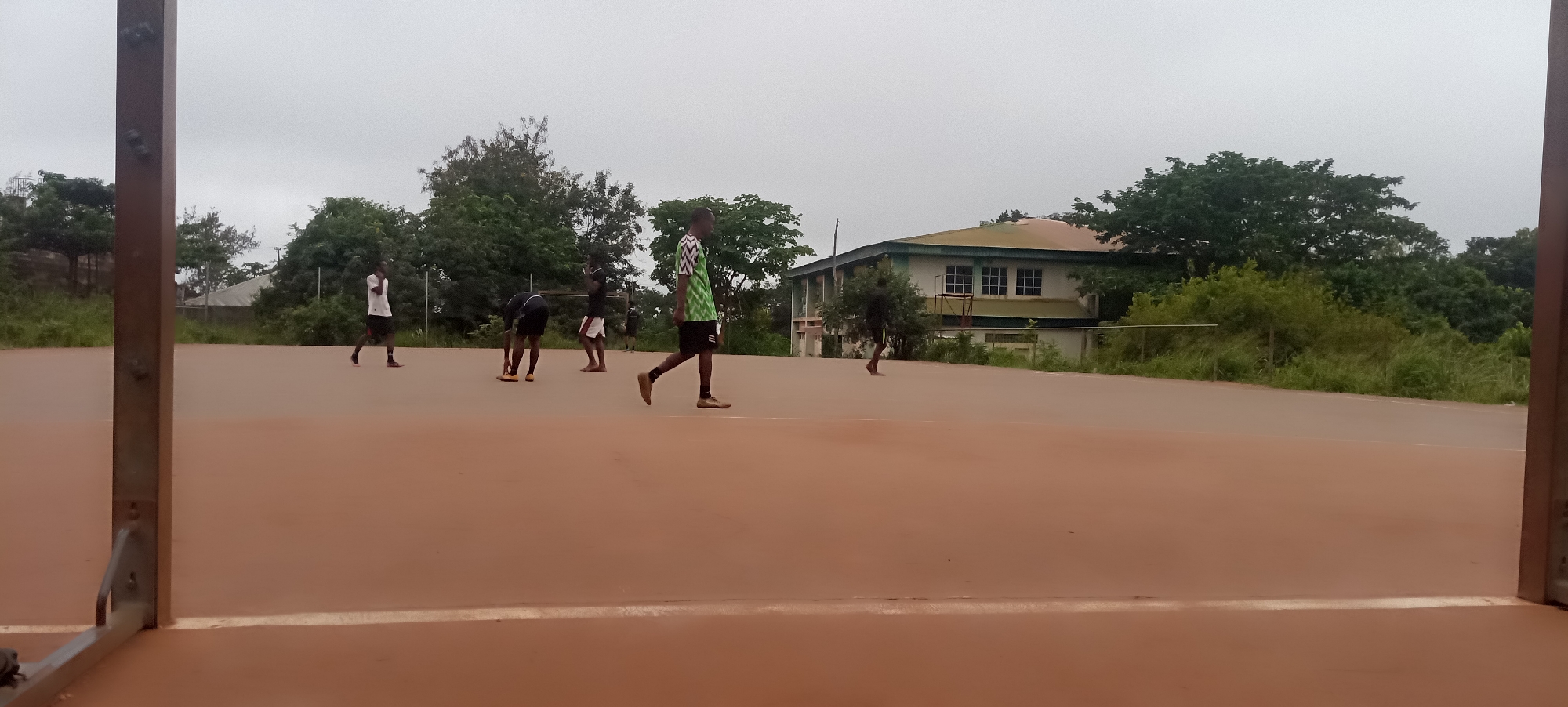 The image depicts an outdoor scene on a reddish-brown court, possibly a sports facility, in Nsukka, Nigeria. The overcast sky suggests it is daytime, possibly late afternoon or early morning. Several young men, dressed in athletic attire, are scattered across the court. One individual in the foreground, wearing a green and white patterned shirt and black shorts, is walking towards the right of the frame. To the left, another person in a white shirt and black shorts is also walking. In the middle ground, several other individuals are present, some appearing to be engaged in a game or practice. One person is bent over, possibly retrieving a ball, while others stand or walk around. In the background, a two-story building with a light-colored facade and a green roof is visible, surrounded by lush green trees and vegetation. A chain-link fence runs along the edge of the court behind the building. The overall impression is of a casual sporting activity taking place in a somewhat unkempt, but functional, outdoor setting.