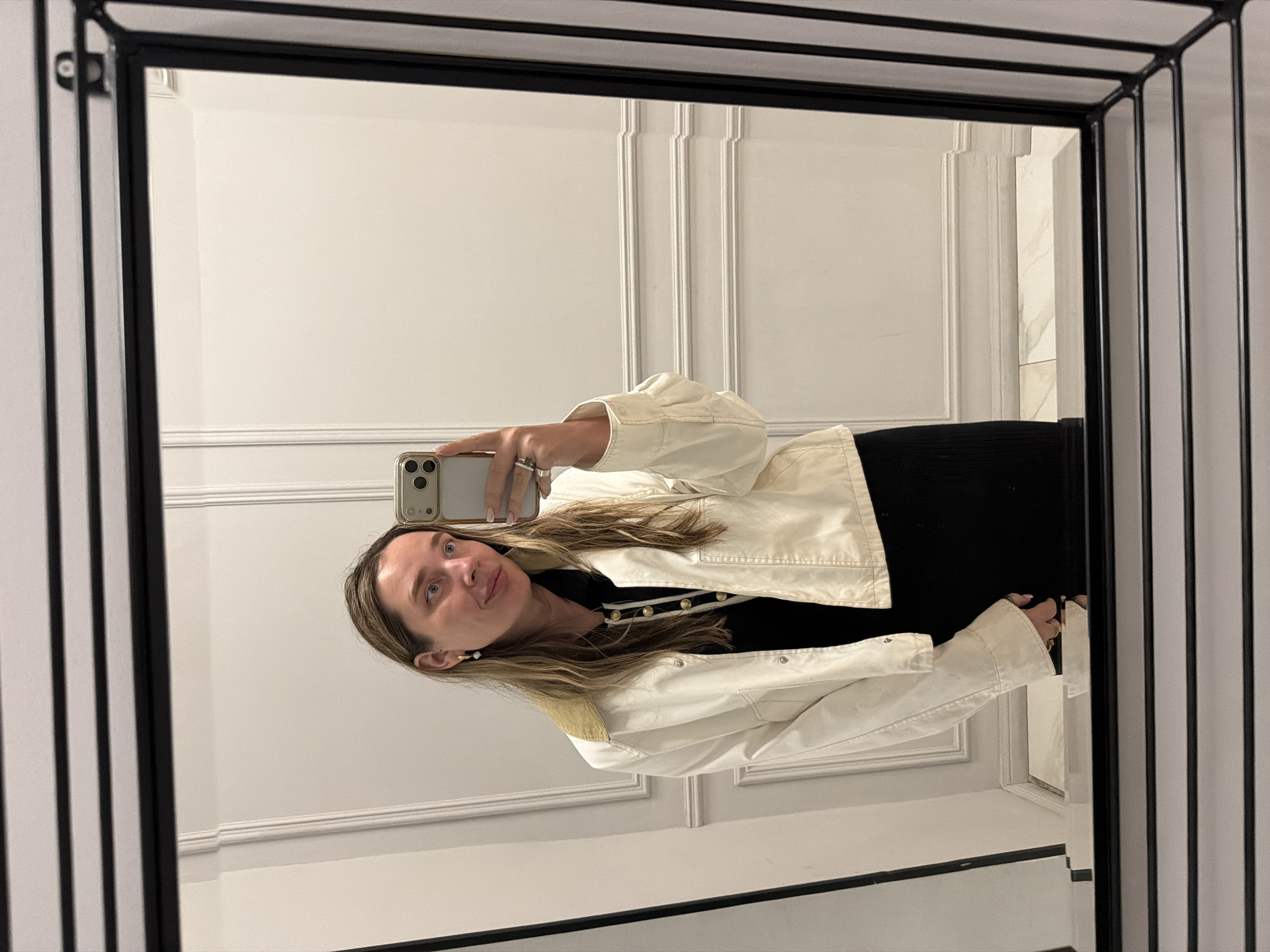 The image is a selfie taken in a mirror. The woman is holding an iPhone in her right hand, and she is wearing a white jacket over a black top. The background is a white wall with decorative moldings. The mirror has a black metal frame. There is no definitive information in the image to indicate the location is Moscow, Russia.