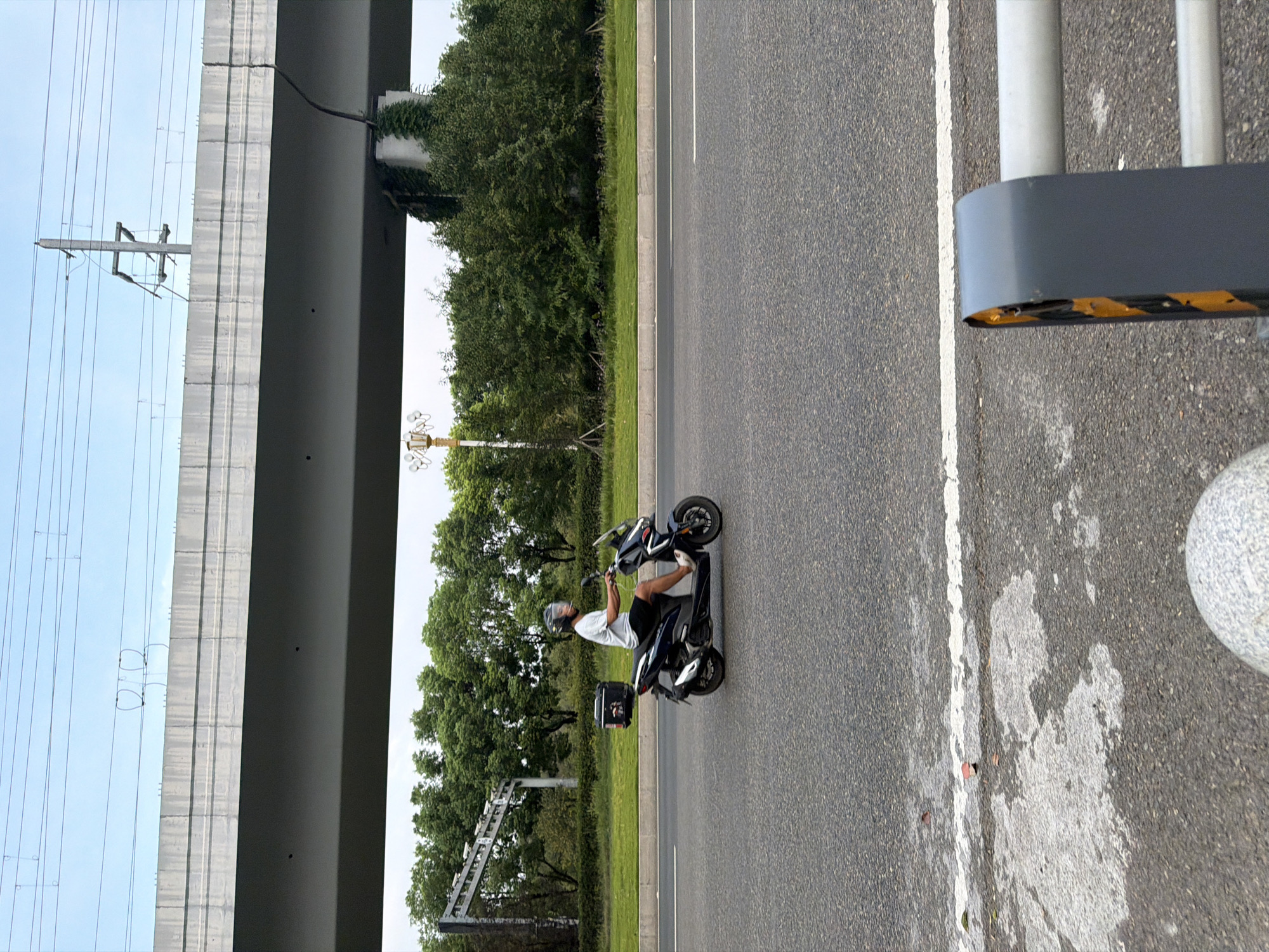 A person wearing a white t-shirt, dark shorts, and a helmet rides a dark-colored scooter on a paved road. The scooter has a black storage box on the back. The road is bordered by a grassy median and trees on the left. In the background, an elevated structure, likely an overpass or railway bridge, spans across the scene, supported by concrete pillars. Overhead power lines are visible above the structure. The sky is overcast. The scene appears to be in the city of Jiangjin, China, based on the provided context. A grey, reflective barrier is partially visible in the foreground on the right side of the image.